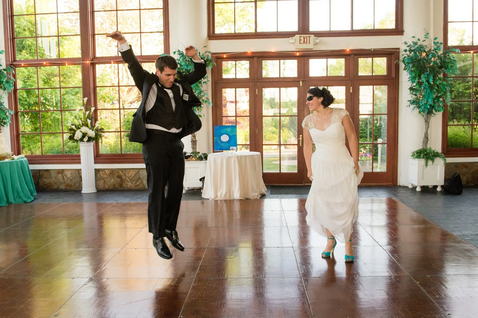 A groom in a black tuxedo jumping with excitement and a bride in a white wedding dress smiling as they dance in an indoor wedding reception hall with large windows and greenery outside.