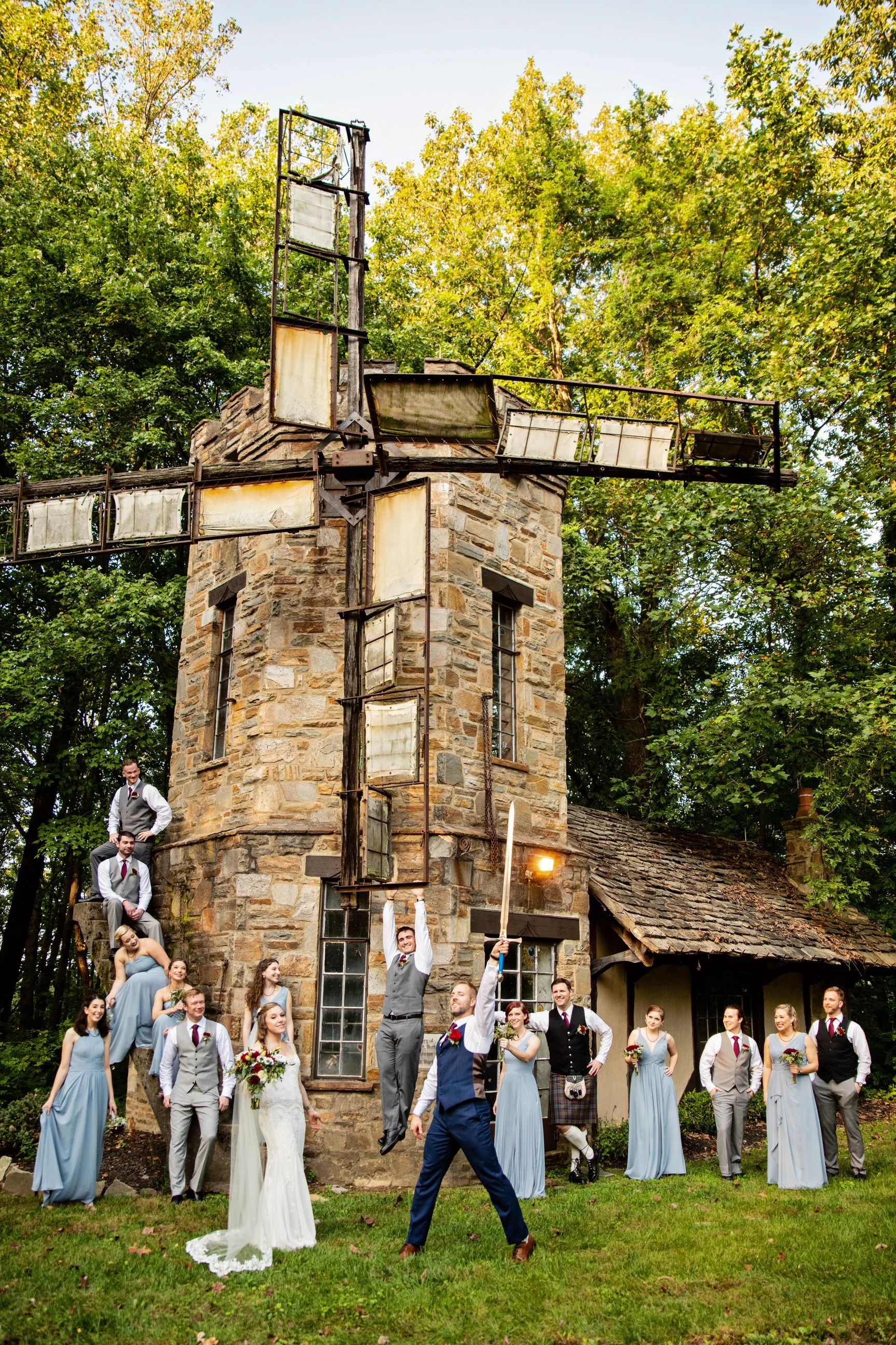 A wedding party in front of a rustic stone windmill, with men in suits and women in bridesmaid dresses, some standing on the ground and some sitting or standing on the windmill's base, celebrating outdoors.