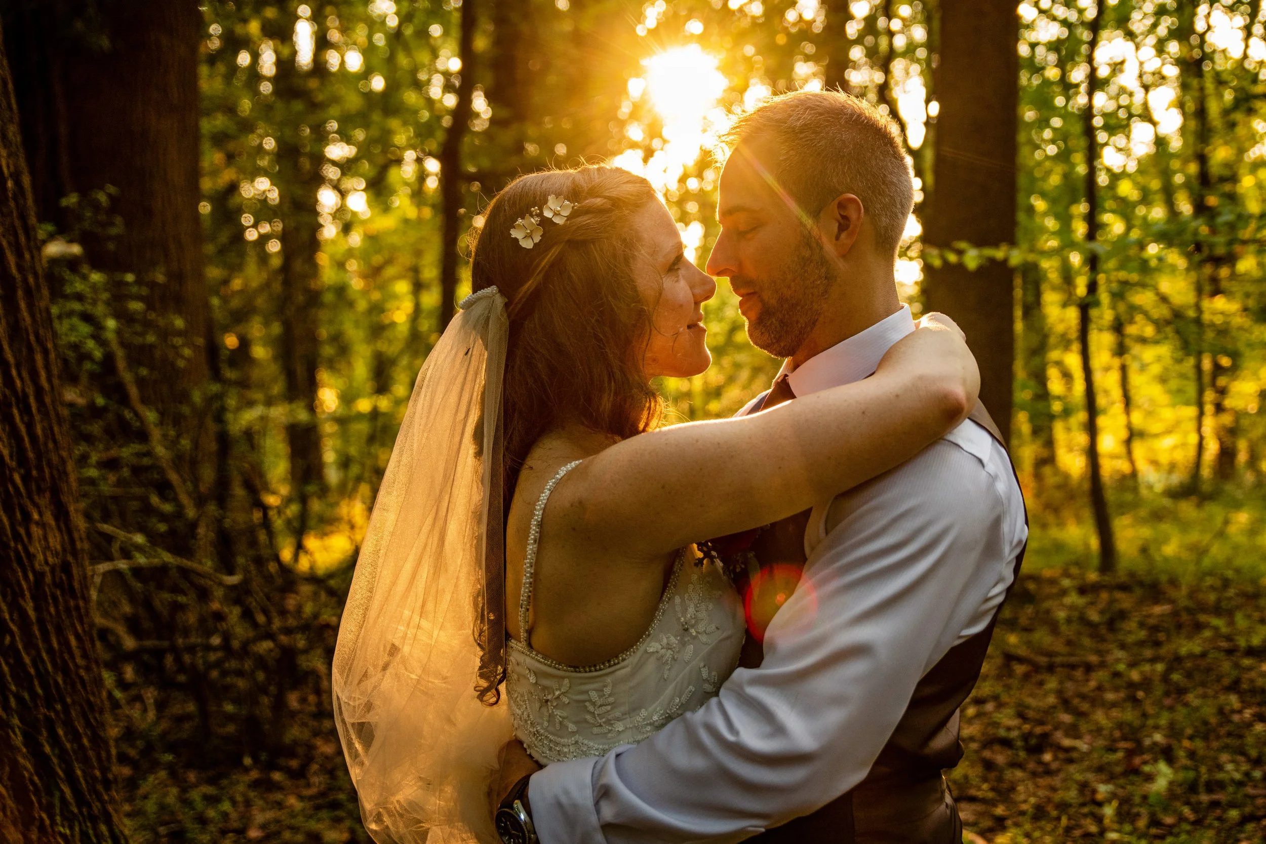 A bride and groom embrace in a sunlit forest at sunset, faces close together, with sunlight filtering through trees in the background.