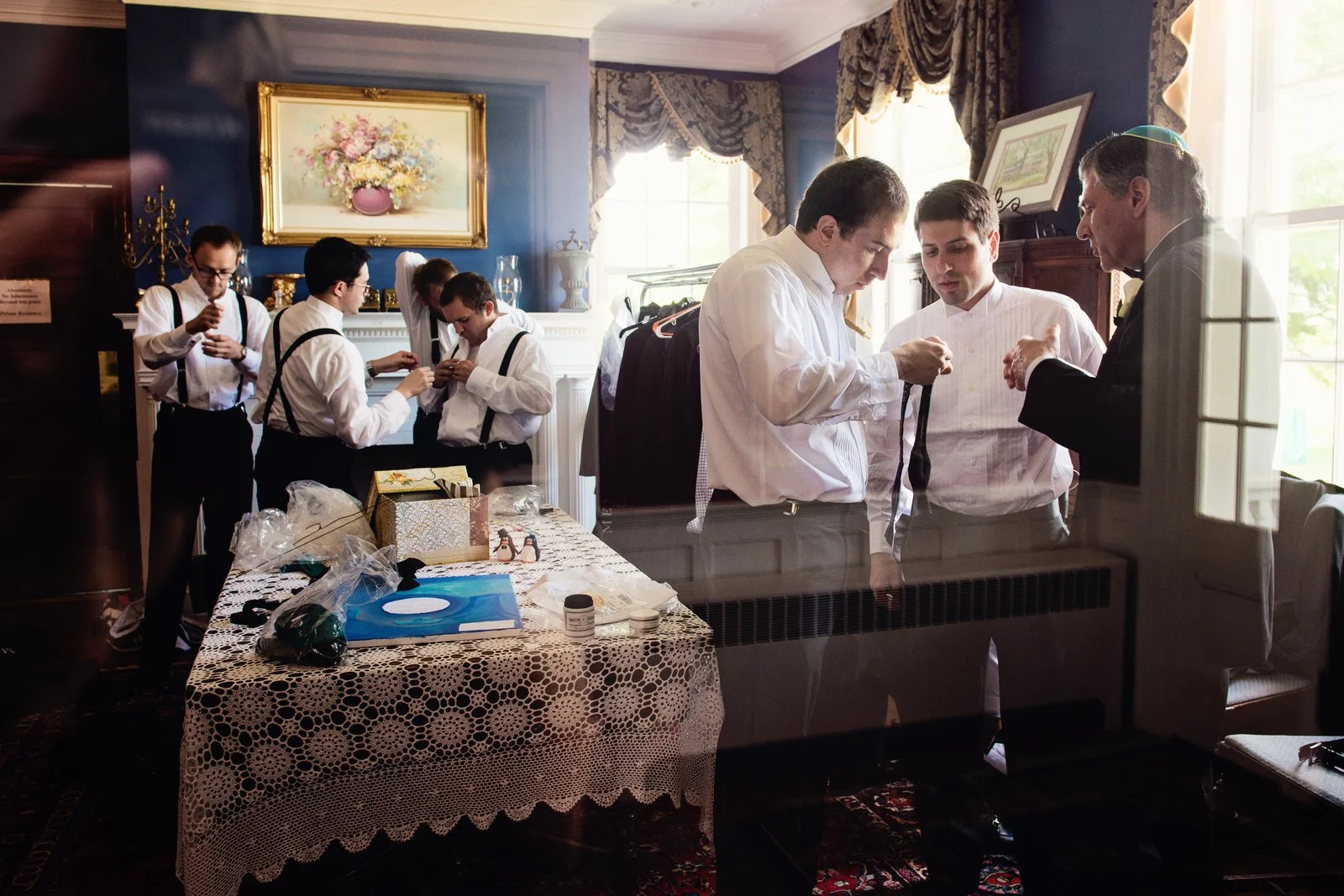 Groom and four men preparing in a well-lit, decorated room with velvet curtains, a table with gifts, and a fireplace.