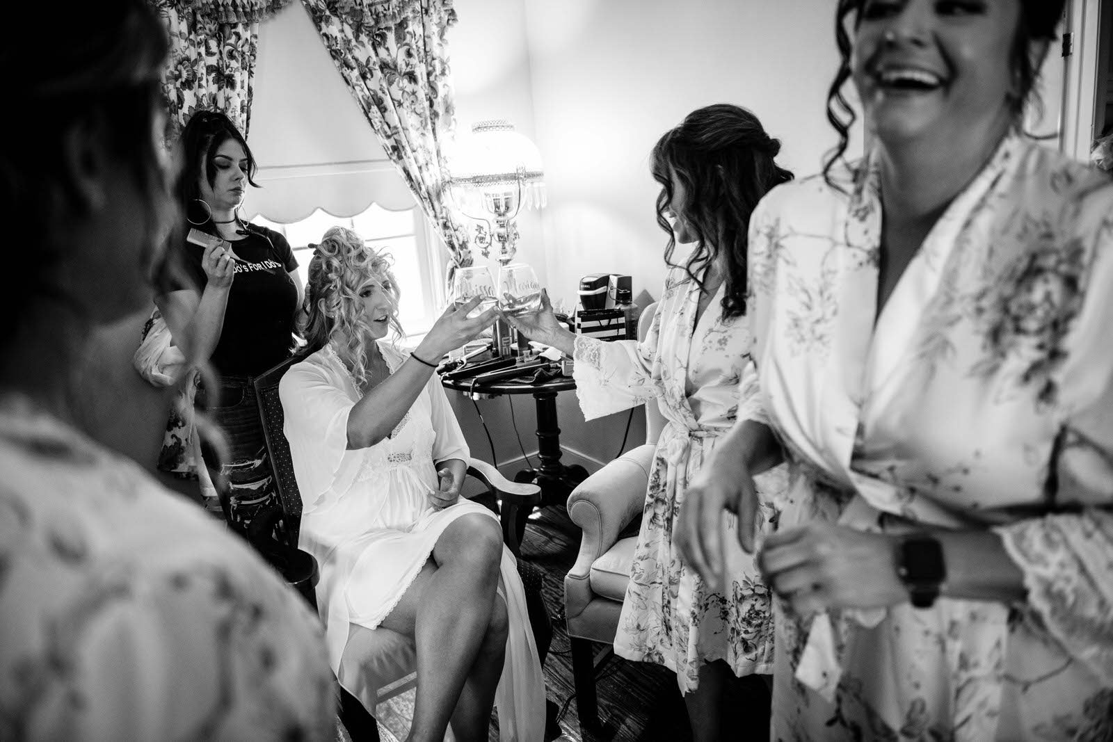 Group of women, with some in floral robes, gather in a room with patterned curtains, toasting glasses in celebration.