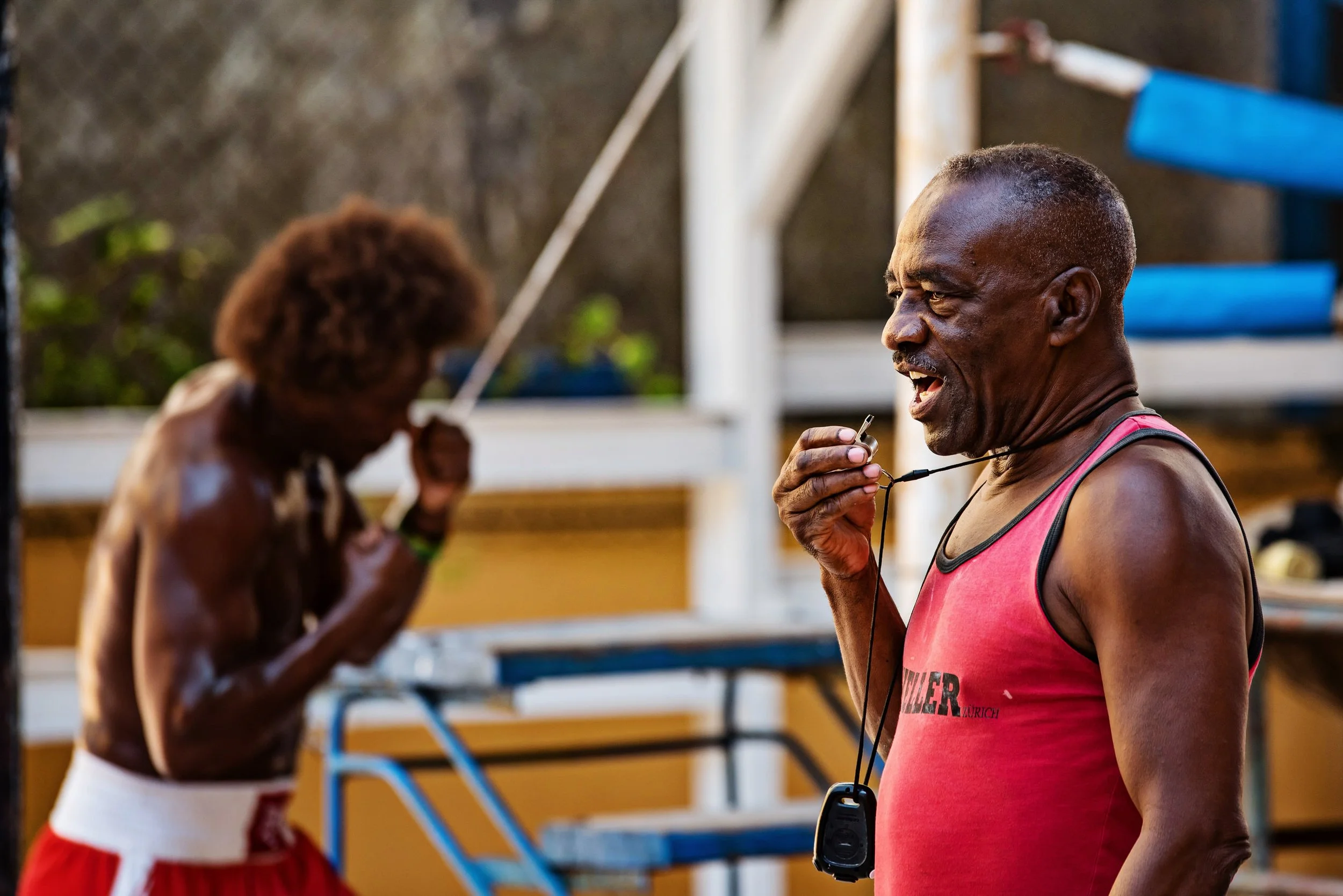 A man in a red tank top talking on a whistle in a boxing gym or outdoor training area with a shirtless man with curly hair in the background adjusting a rope.