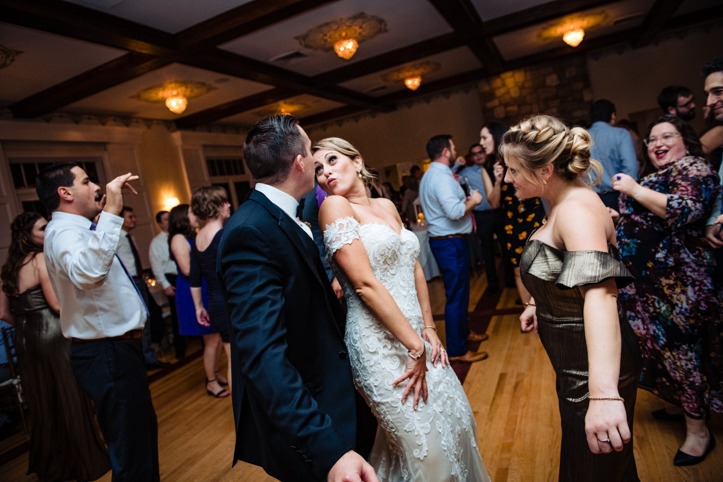 A wedding reception with guests dancing and socializing, a woman in a white lace wedding dress, and a man in a black tuxedo in the center.