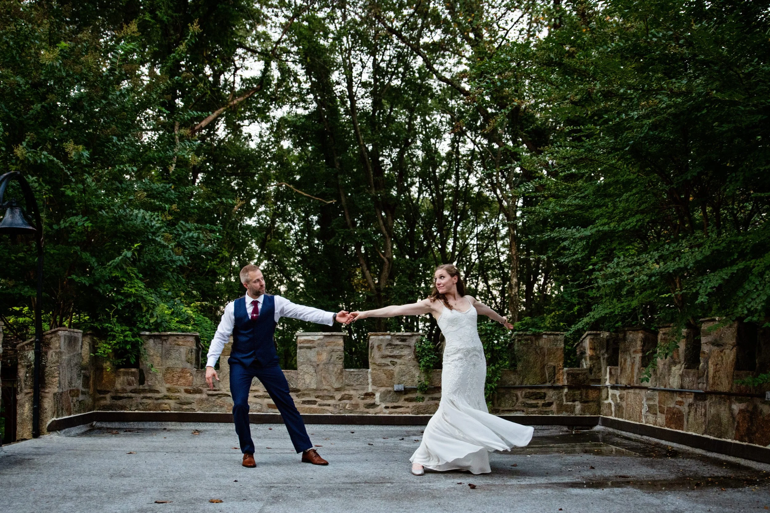 A man and woman dancing outdoors, woman in a white wedding dress and man in a navy suit, on a paved area with stone wall and lush green trees in the background.