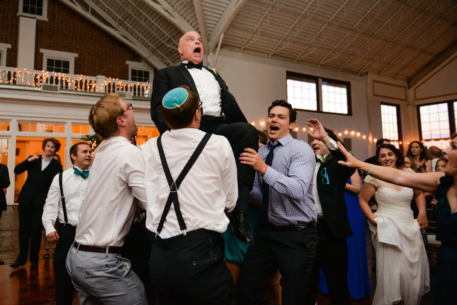 Group of people celebrating at a wedding reception as they lift a man in a tuxedo into the air.