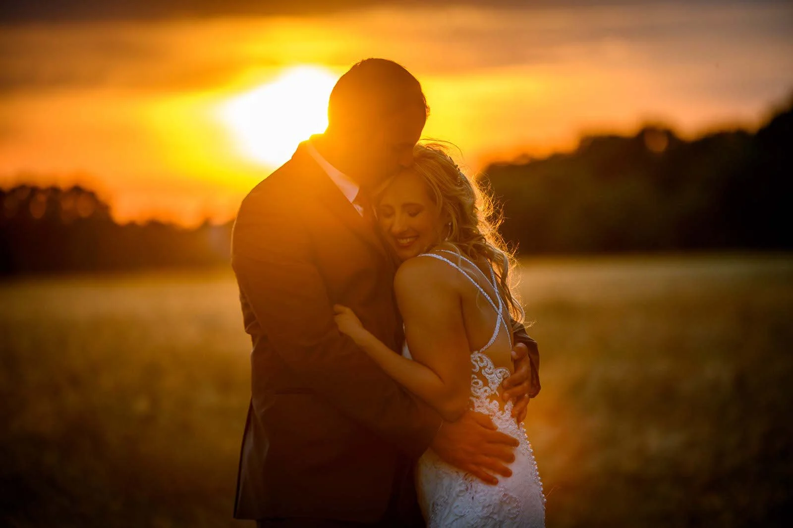 A couple hugging at sunset, with the sun setting in the background. The woman is wearing a white lace dress, and the man is wearing a brown suit. They look happy and tender, embracing each other closely.