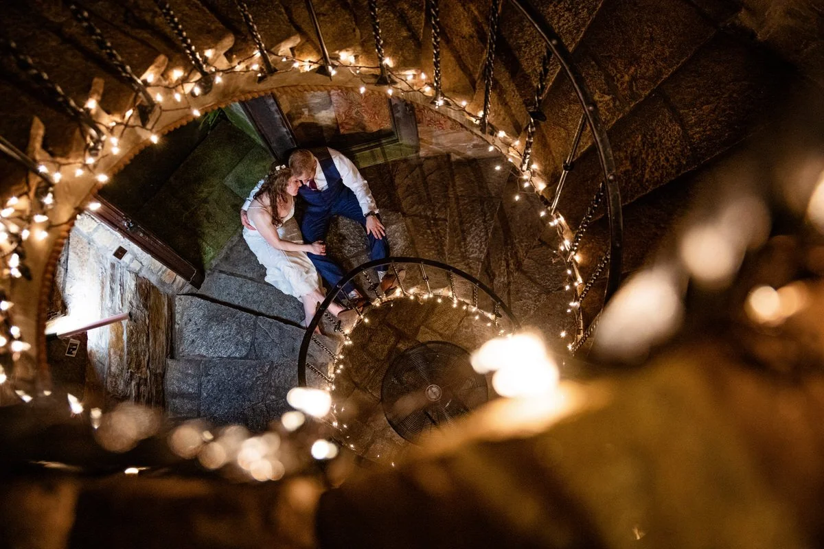 A couple sitting on stone stairs at the bottom of a spiral staircase decorated with string lights.