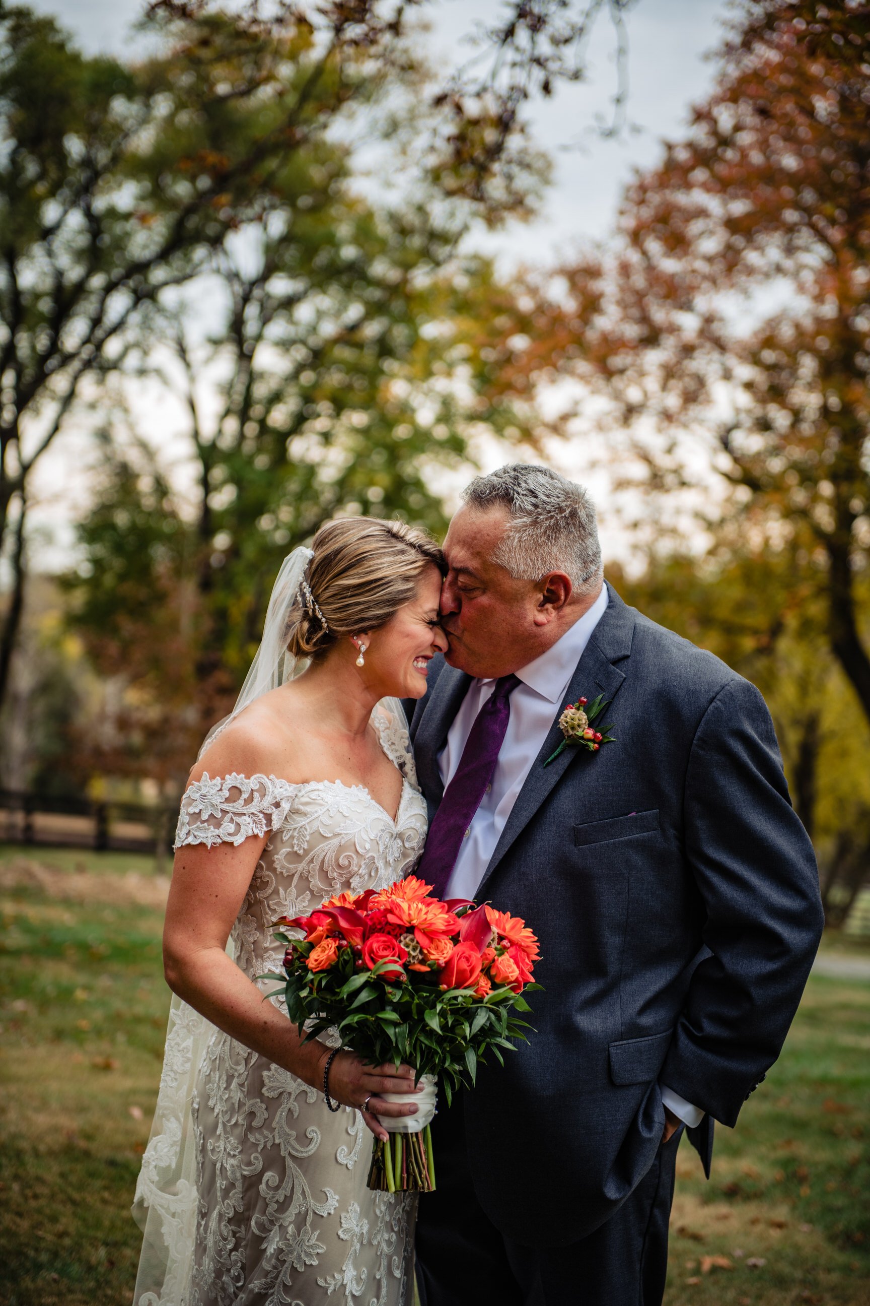 A bride and an older man, likely her father, share a tender moment outdoors during a wedding, touching foreheads with eyes closed, the bride holding a bouquet of colorful flowers, surrounded by trees with autumn leaves.