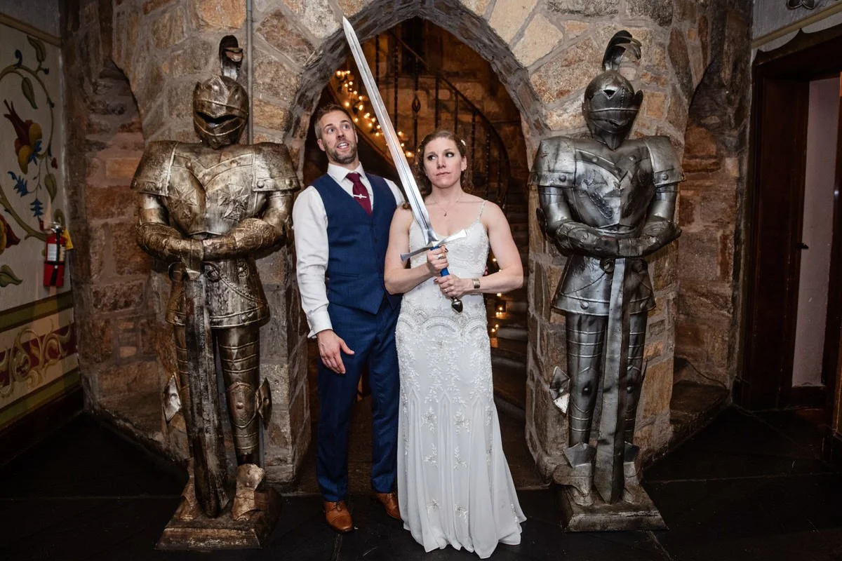 A man in a suit and tie and a woman in a wedding dress holding a large sword pose between two suits of medieval armor statues in a rustic stone setting.
