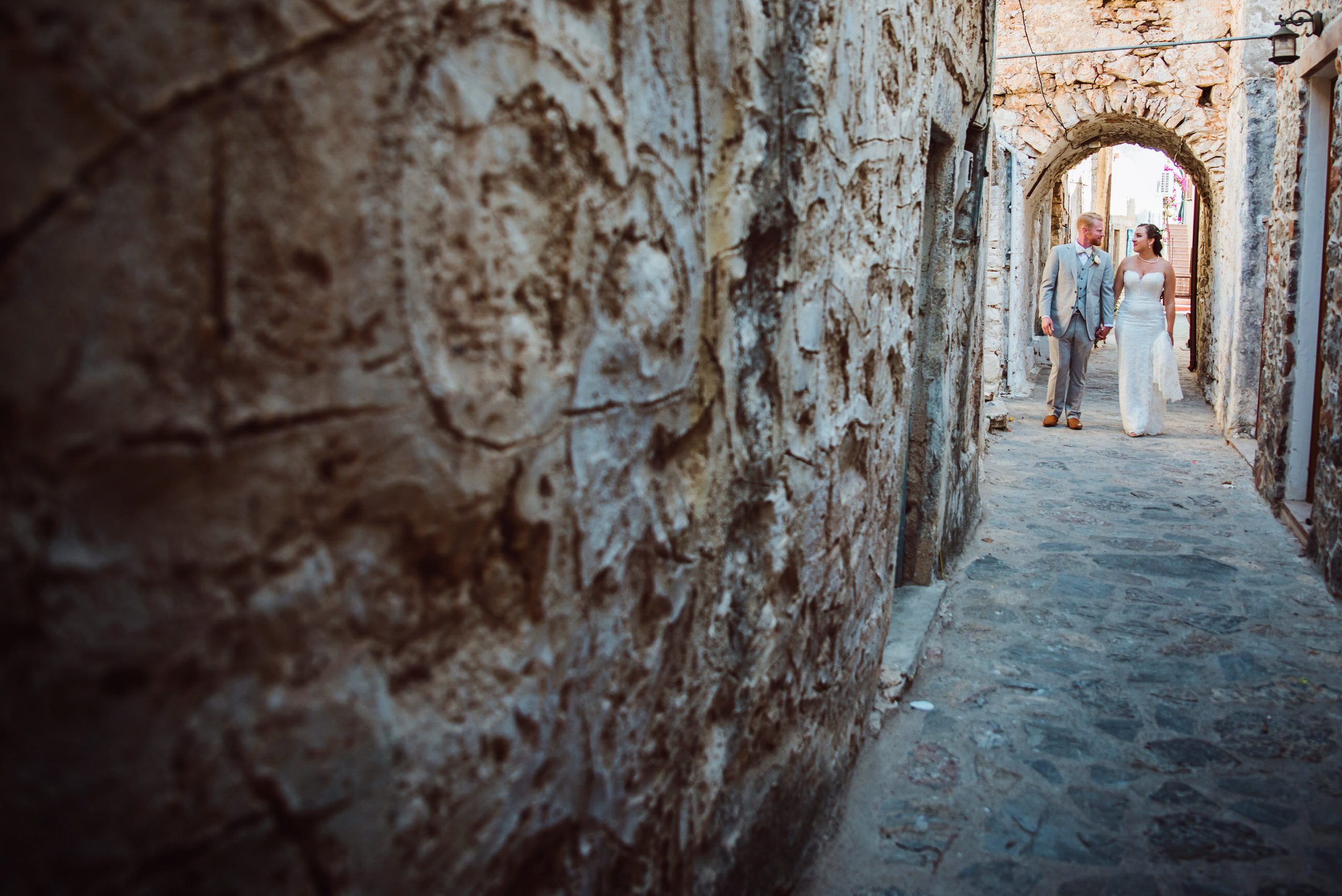 A couple dressed in wedding attire holding hands and walking through a narrow, stone-arched alleyway with textured stone walls.