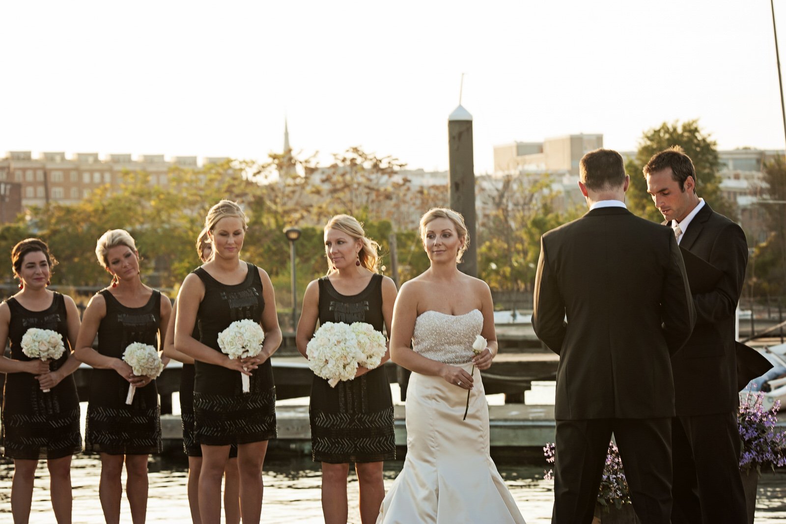 A wedding ceremony with the bride, groom, bridesmaids, and officiant outdoors near a body of water, with autumn trees and city buildings in the background.