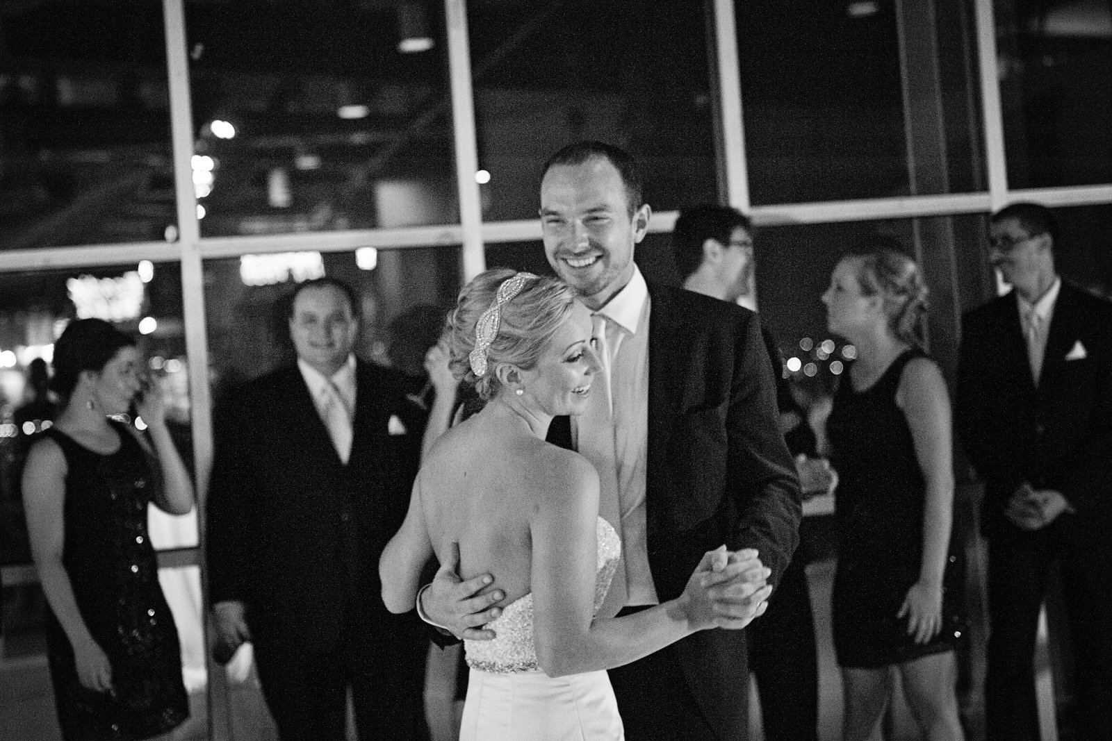 A bride and groom sharing a dance at their wedding reception, surrounded by wedding guests in formal attire, in a dimly lit venue.