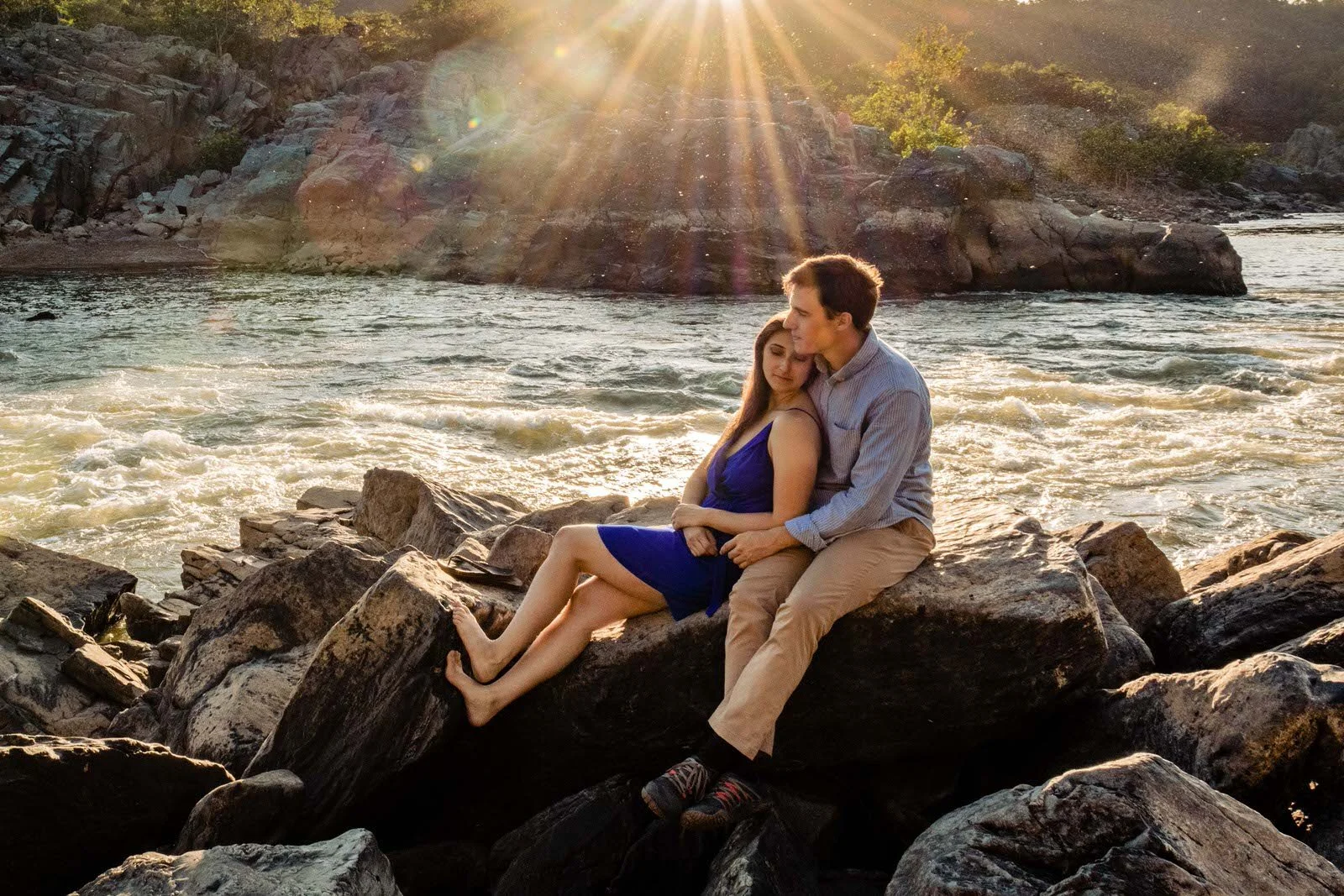 A young couple sitting on rocks by a river at sunset, embracing each other. The woman is in a blue dress, and the man is in a light-colored shirt and khakis. The sun is low in the sky, casting warm light and rays over the scene.