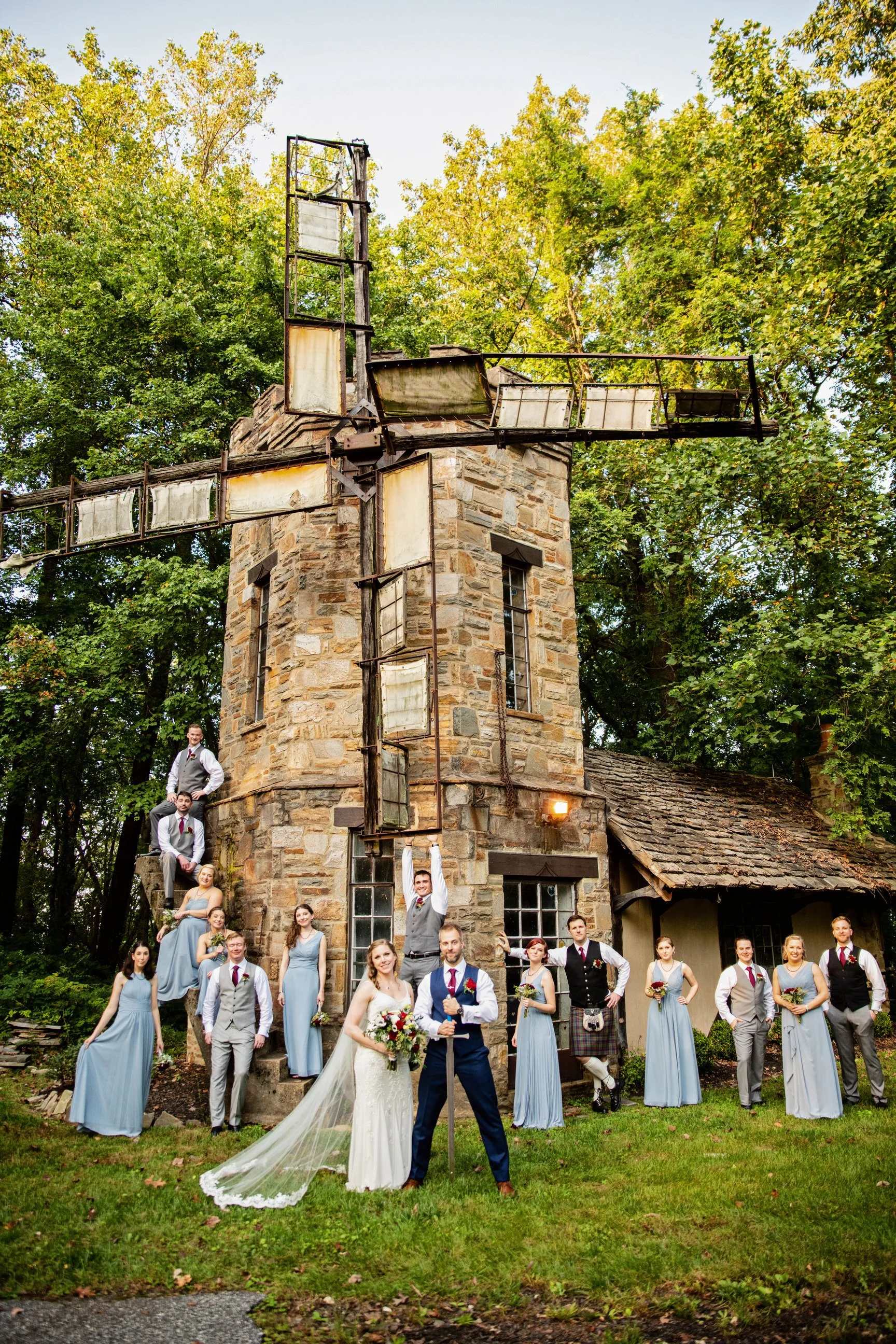 A wedding party stands in front of a rustic stone windmill, with bridesmaids in light blue dresses and groomsmen in suits, outside amidst trees.