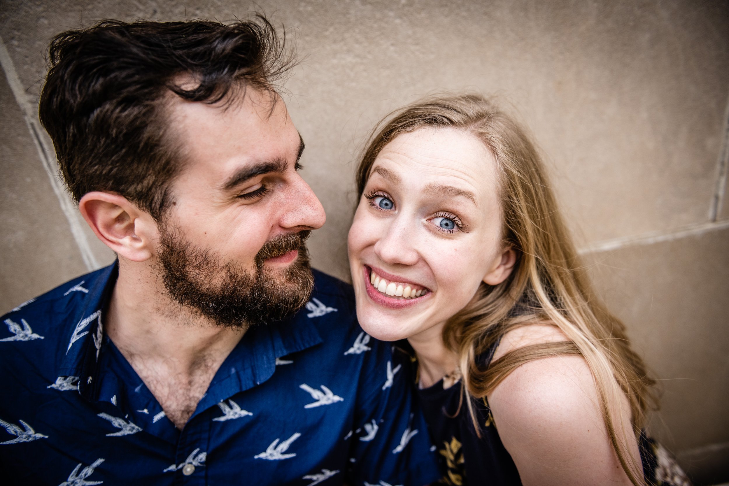 A smiling woman with blue eyes and long blonde hair leaning close to a bearded man with dark hair, both looking happy and happy.