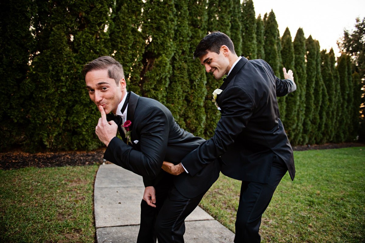 Two men dressed in tuxedos, one behind the other on a sidewalk, with one holding the other's waist, both smiling in a humorous pose outdoors with green trees in the background.