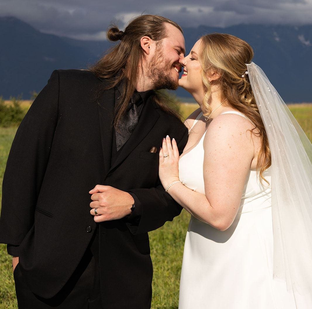 Outdoor Montana wedding couple portrait — bride in white gown and veil with groom in black suit laughing together in a green field against dramatic mountain and storm cloud backdrop.