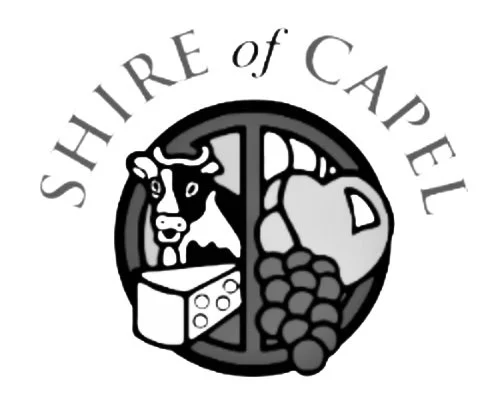 A back and white image that reads the shire of capel, the image has a cow, cheese, grapes and greenery inside a circular window frame.