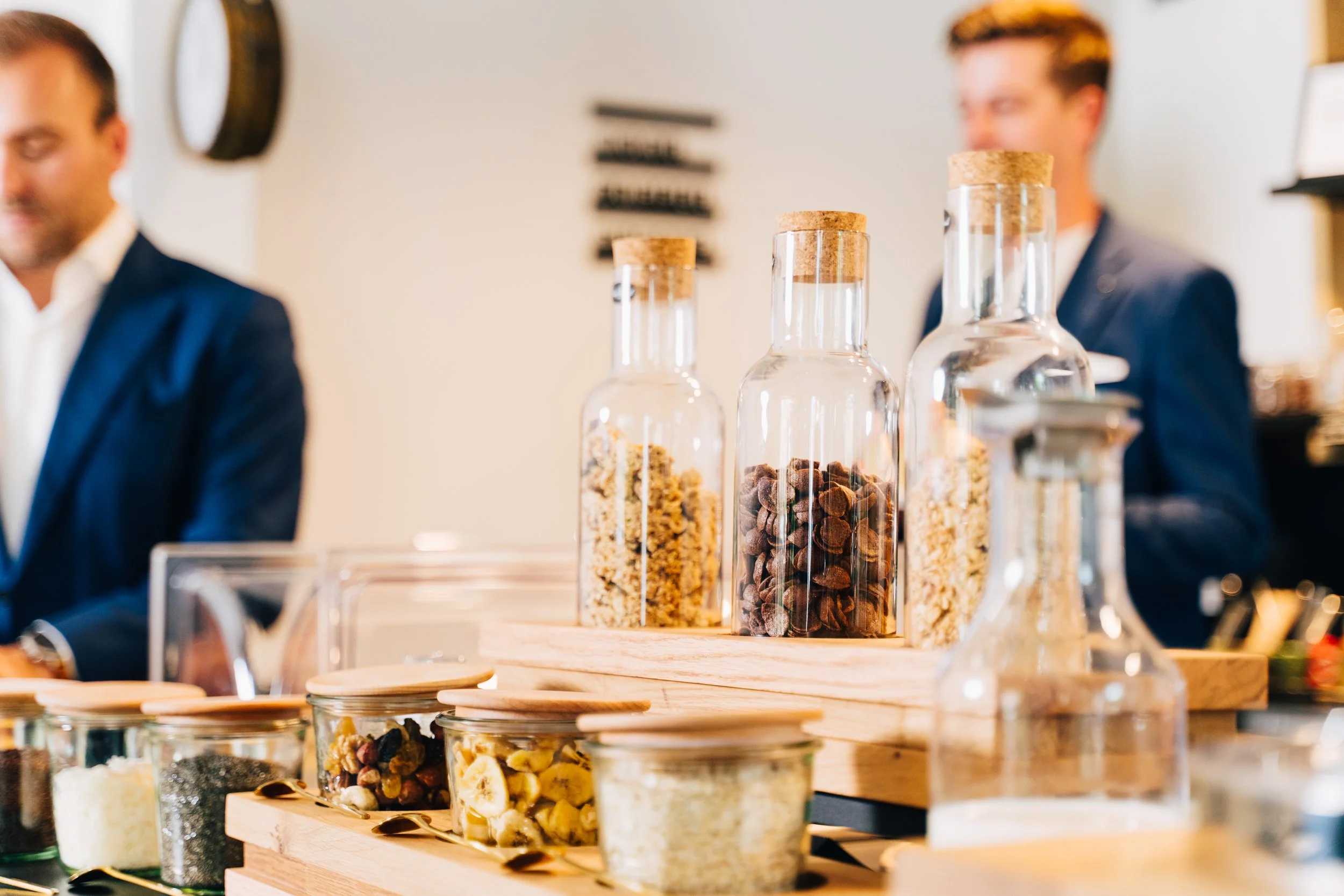 Verschiedene Gläser und kleine Behälter mit Nüssen, Samen und getrockneten Früchten auf einem hölzernen Präsentierteller, im Hintergrund zwei Männer in ordentlichen Jacken.