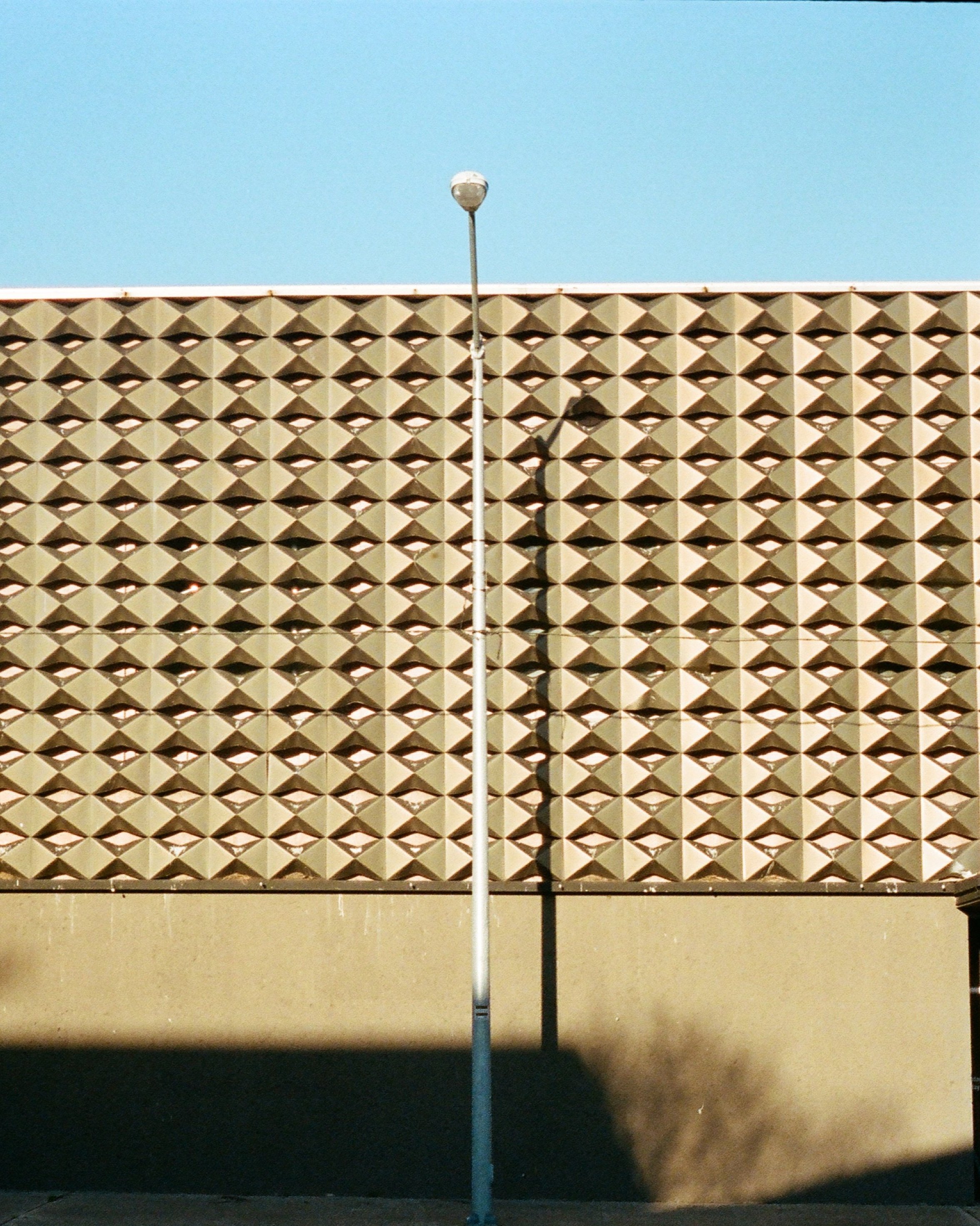 A beige building with a textured, geometric pattern of diamond-shaped openings, a tall street lamp in front of it, and shadow cast on the lower part of the wall.