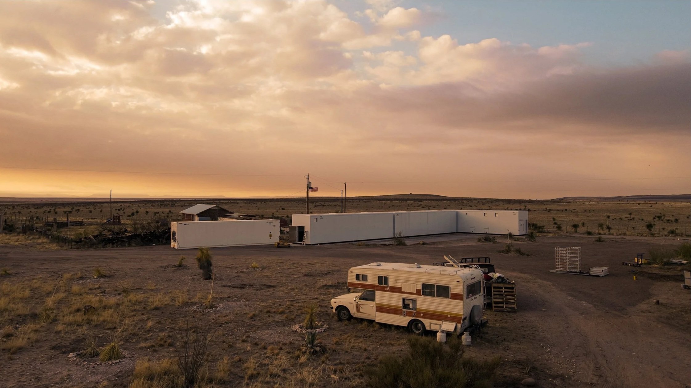 A desert landscape at sunset with an RV parked on dirt ground, a white modular building, and sparse vegetation in the background.