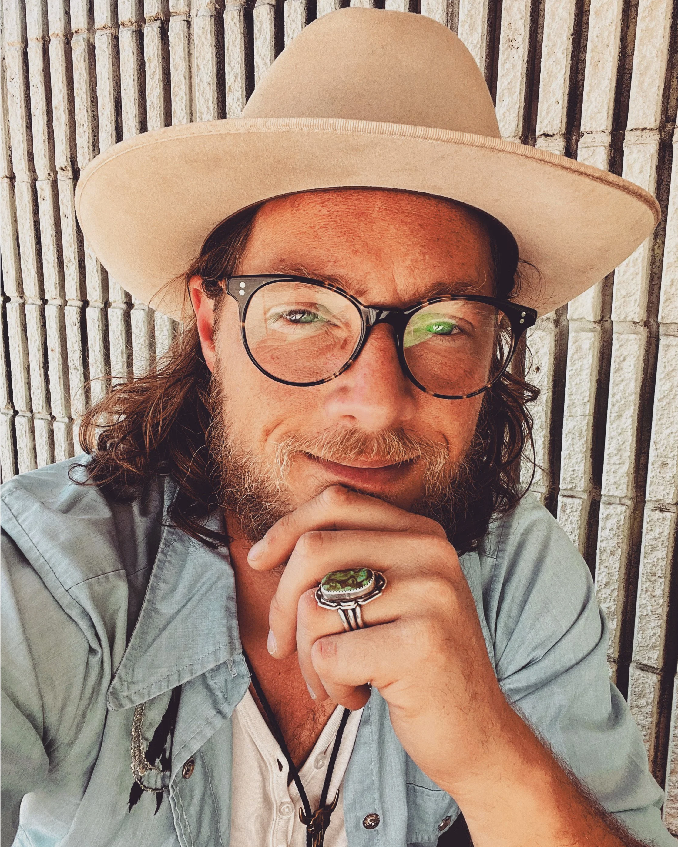 A man with glasses, a light-colored hat, and long hair, smiling with his hand resting on his chin in front of a brick wall.