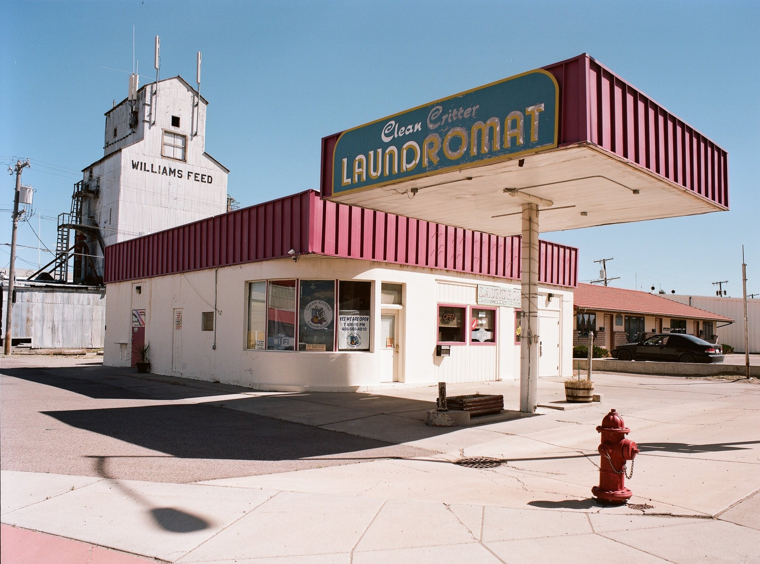 A vintage laundromat with a large sign reading "Clean Critter Laundry" in a small town. The building has white walls with a pink roof and pink trim, and a window with a success message and Wi-Fi sign. A red fire hydrant is in the foreground, and there are neighboring houses and a grain elevator with "Williams Feed" written on it in the background.