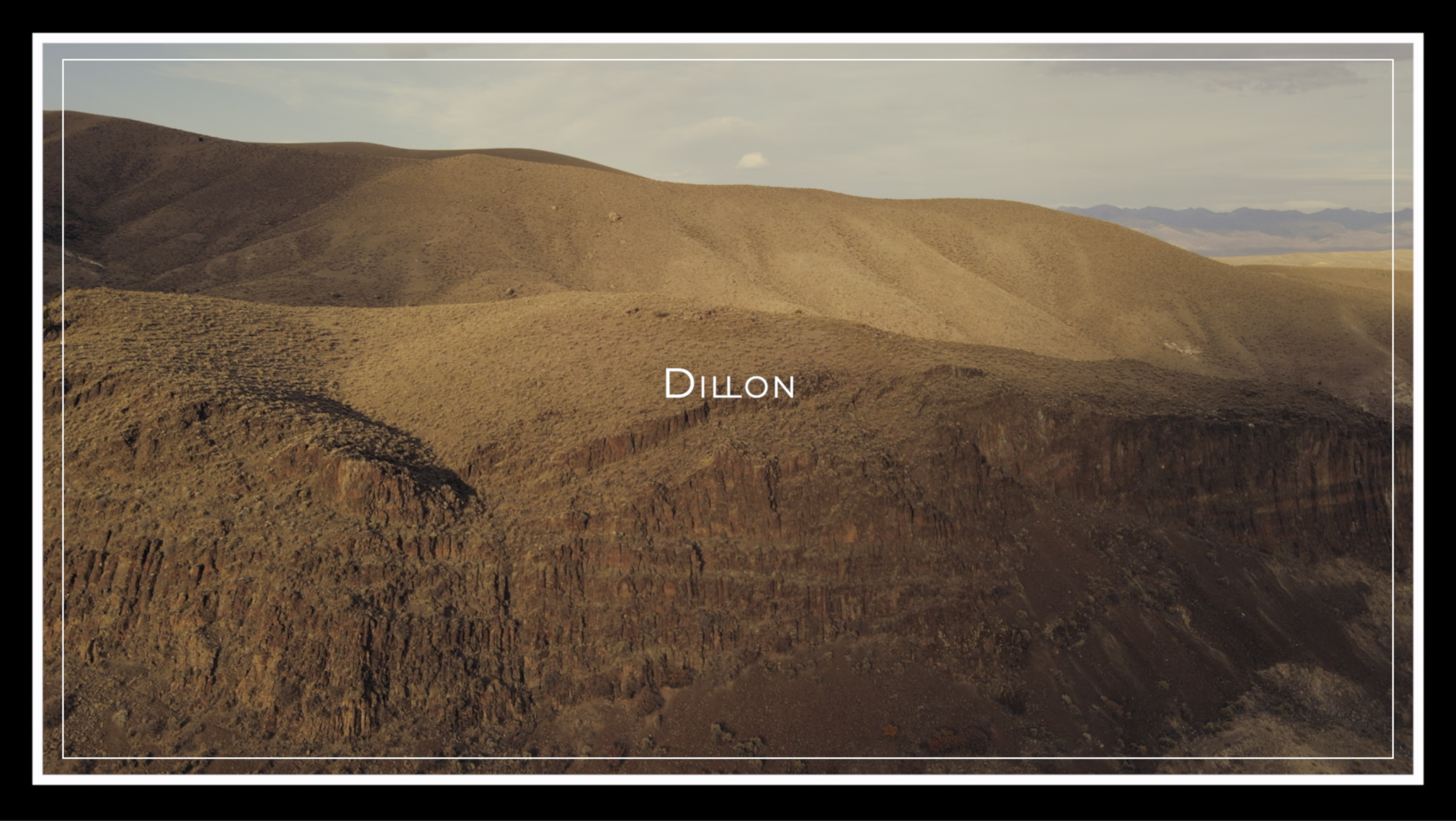 Aerial view of a rugged, arid hillside in Dillon, with dry, brown grass and a steep clay cliff in the foreground under a partly cloudy sky.