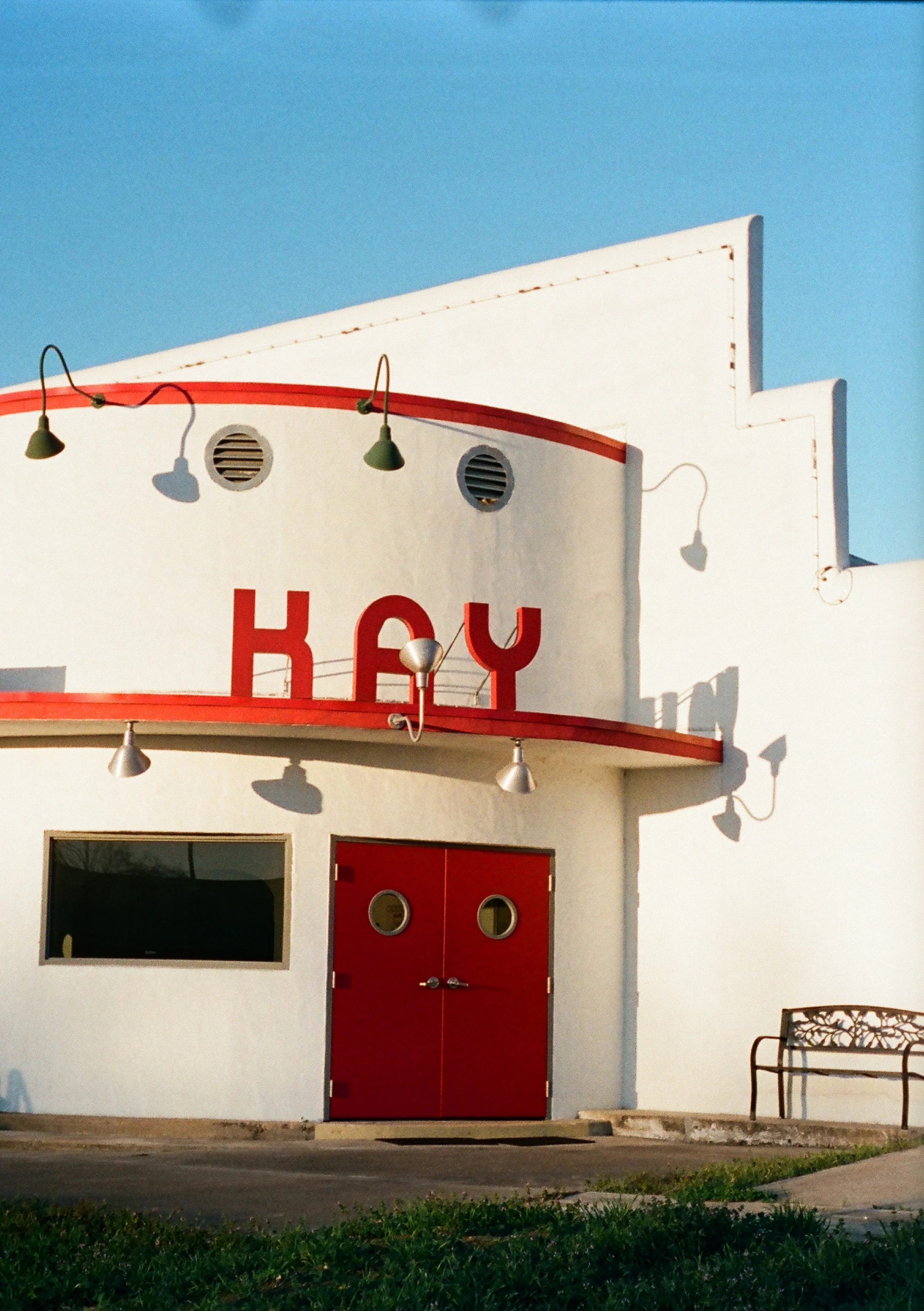 A white building with red accents and large red letters spellingK+Y, featuring a pair of red double doors with round windows, a portrait window, and a bench outside on a sunny day.
