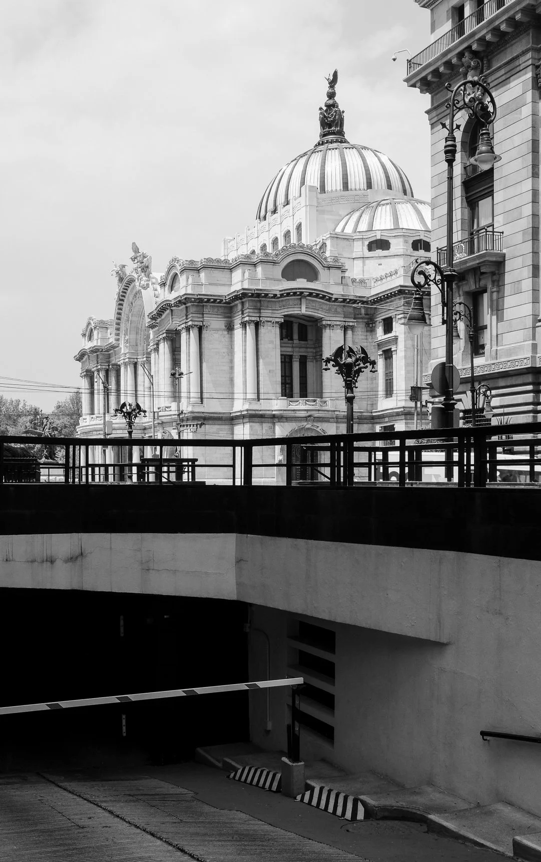Black and white photograph of a historic domed building with classical architecture, ornate details, and sculptures on the roof. The building is viewed from a lower level with street lamps and a parking garage entrance in the foreground.