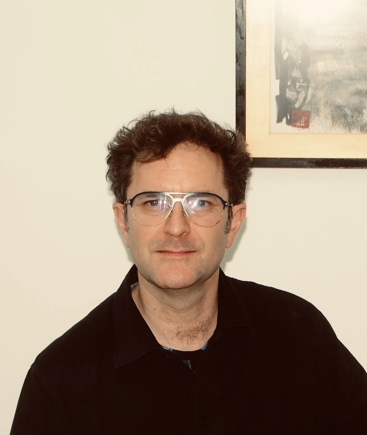A man with short dark hair, wearing glasses with a metal frame, and a black shirt, standing in front of a plain light-colored wall with a framed picture hanging behind him.