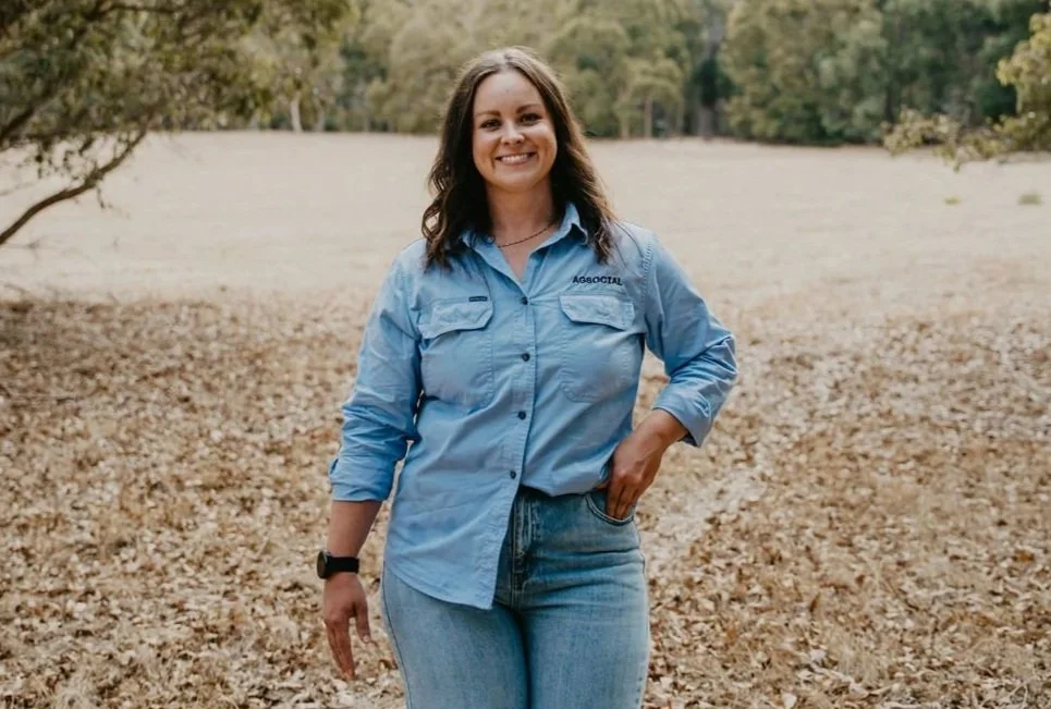 Woman with brown hair smiling, wearing a light blue long-sleeve shirt and jeans, standing outdoors in a wooded area with dry leaves on the ground and trees in the background — representing AGSOCIAL’s grounded and regionally connected approach to agri