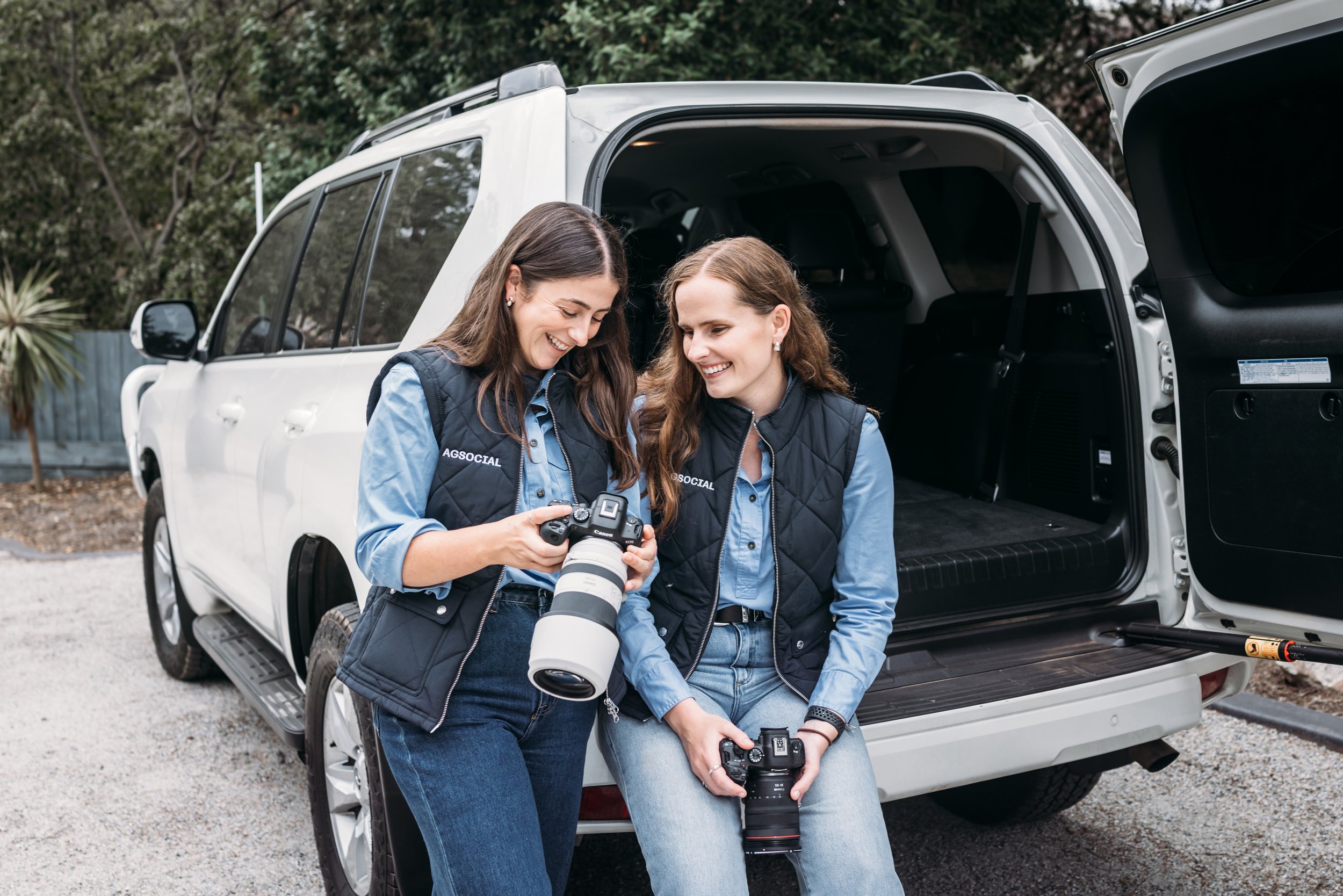 Woman with brown hair in a ponytail filming two people walking across a dry rural field using a digital camera, with scattered trees and a partly cloudy sky — showcasing AGSOCIAL’s on-farm content creation for agricultural brands.