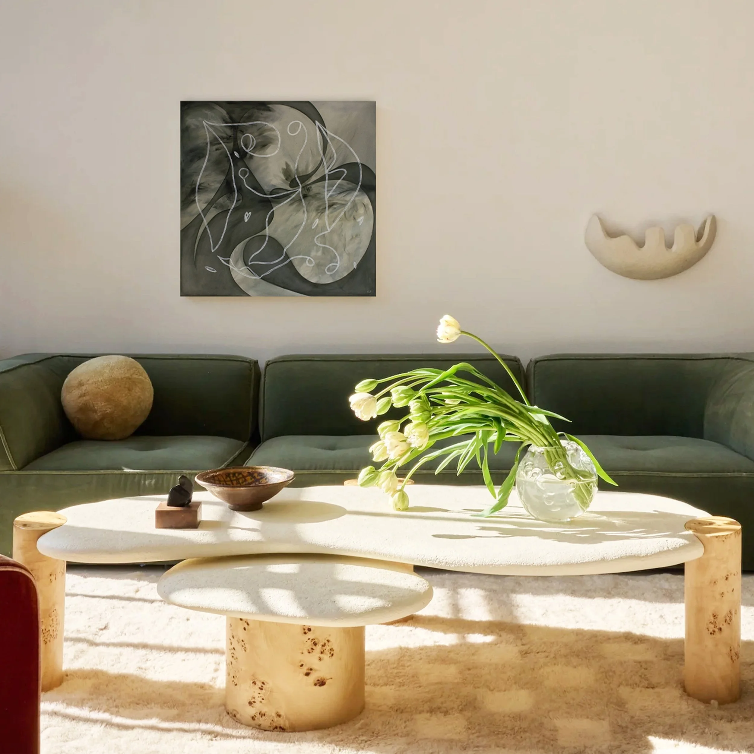 Living room with a green sofa, abstract wall art by Laura Wickstead, a beige wall-mounted sculpture, a white textured coffee table with a glass vase of white flowers, and decorative objects.