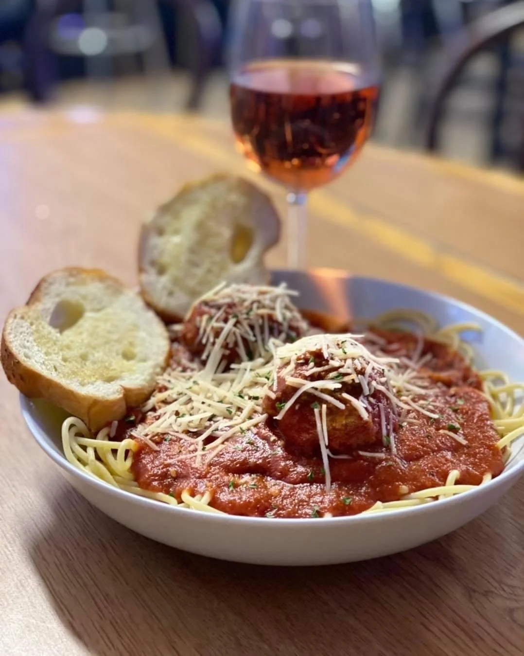 A bowl of spaghetti with tomato sauce, topped with grated cheese, served with slices of bread and a glass of rosé wine in the background.
