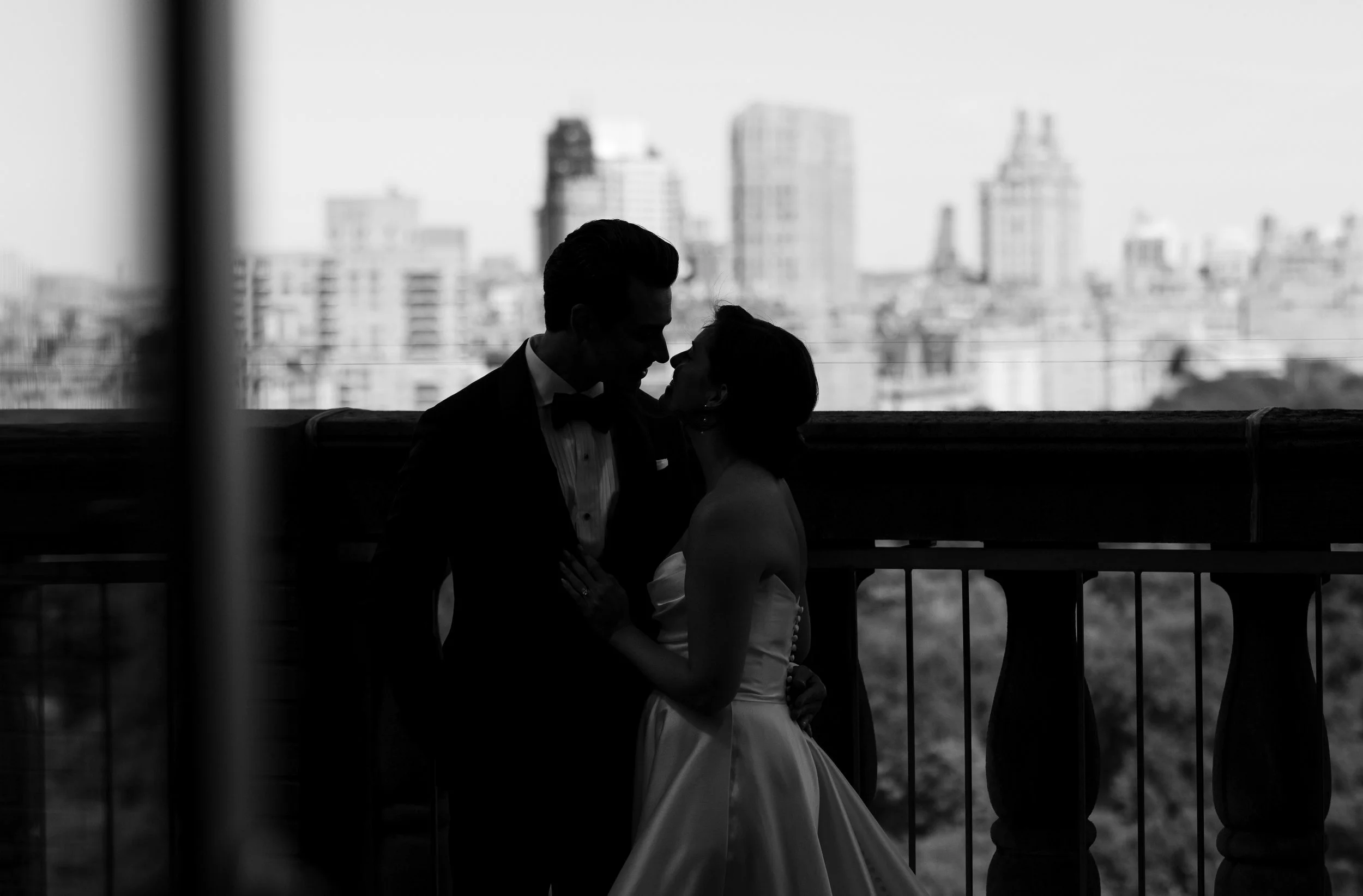 Silhouetted couple in wedding attire kissing on a balcony with city skyline in the background.