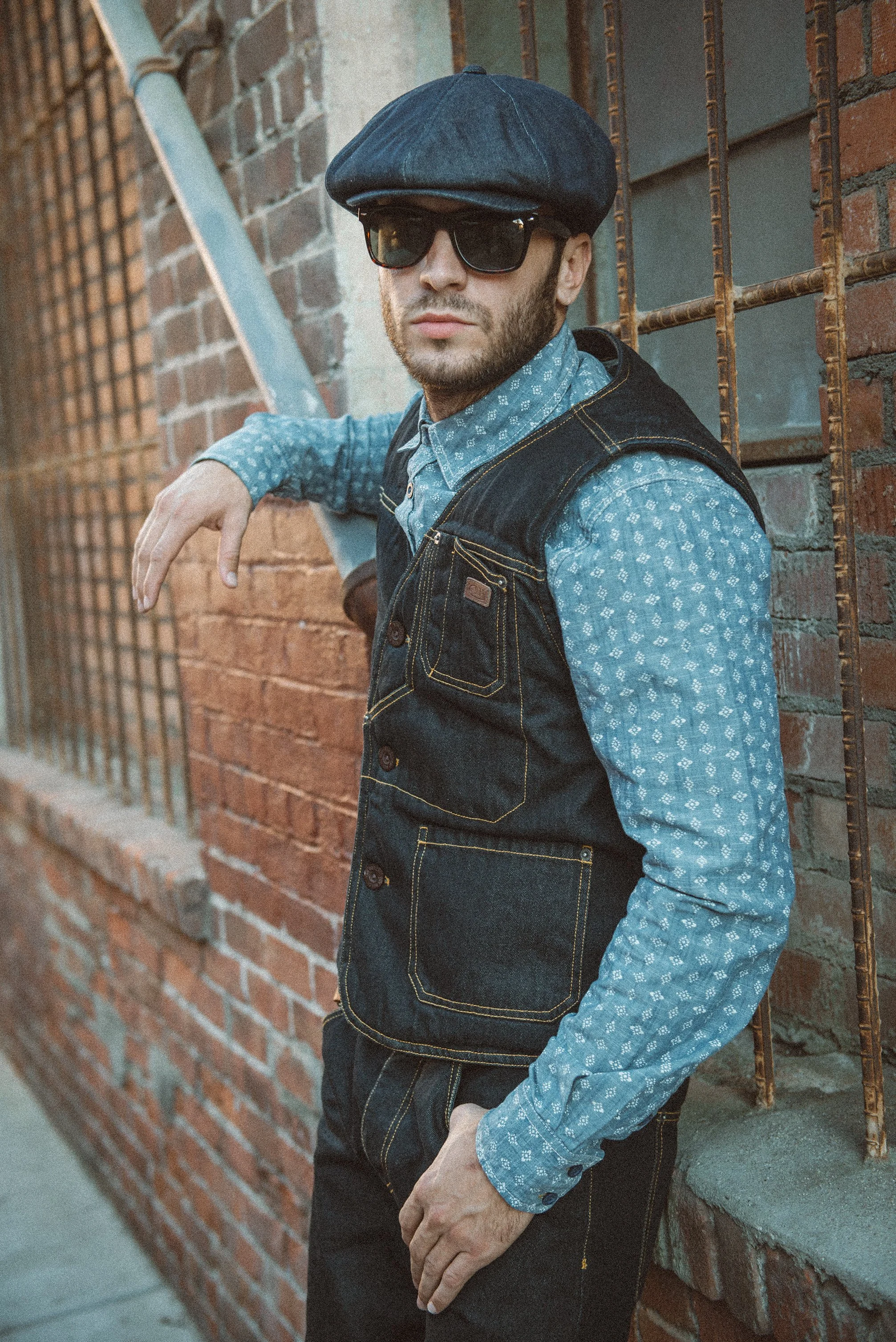 A man wearing a denim flat cap, black sunglasses, a blue patterned shirt, and a dark denim vest leaning against a brick wall with one hand on his hip.