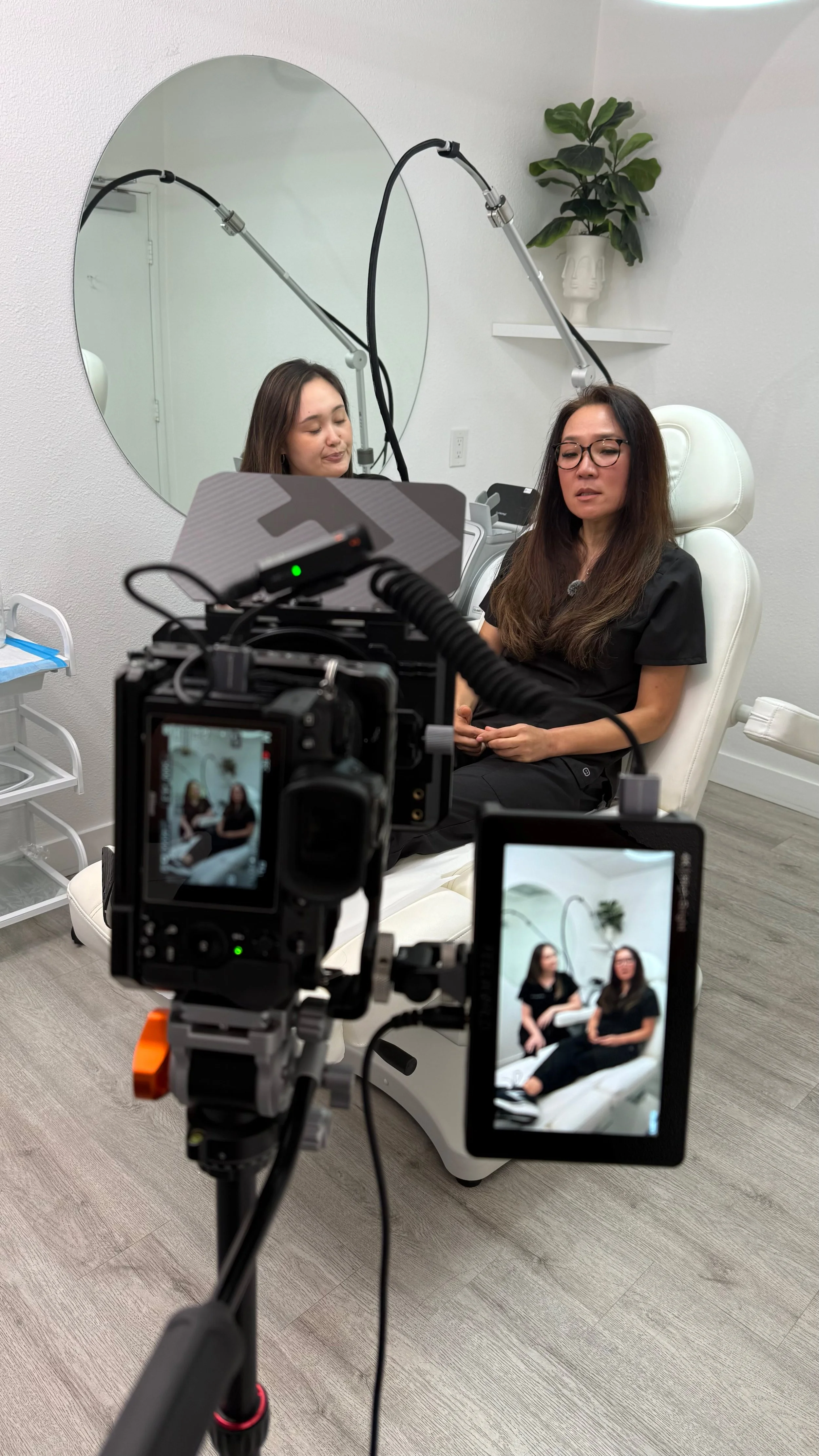 Two women are sitting in a medical office, being recorded by a camera on a tripod. One woman has long brown hair, wears glasses, and a black shirt. The other woman has shoulder-length brown hair and is seated with closed eyes. A large mirror is on the wall behind them, and a potted plant is on a shelf above them.