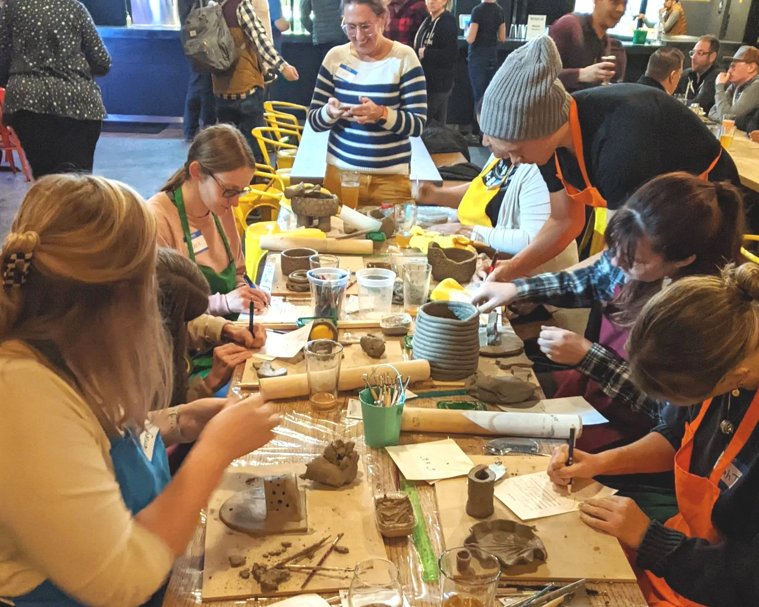 Group of people gathered around a table engaging in arts and crafts, working with pottery and clay sculptures, in a cozy indoor setting.