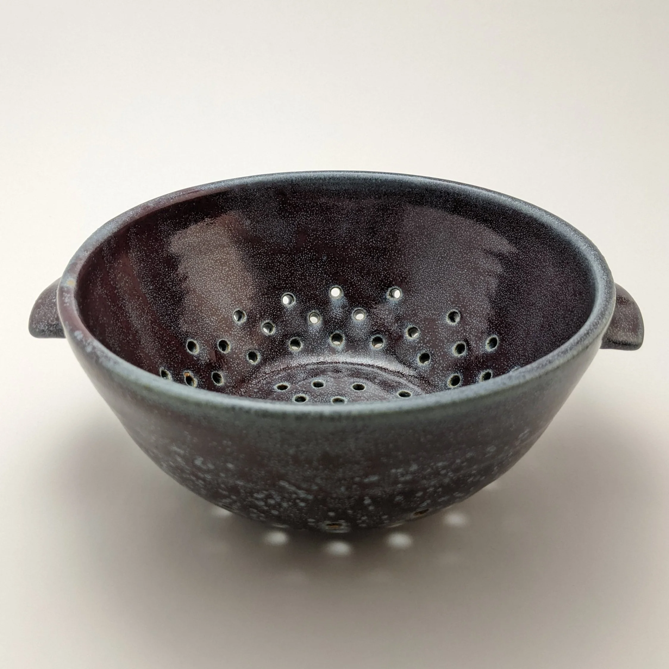 Close-up of a dark, textured ceramic bowl with small holes, placed on a white surface with a plain light background.