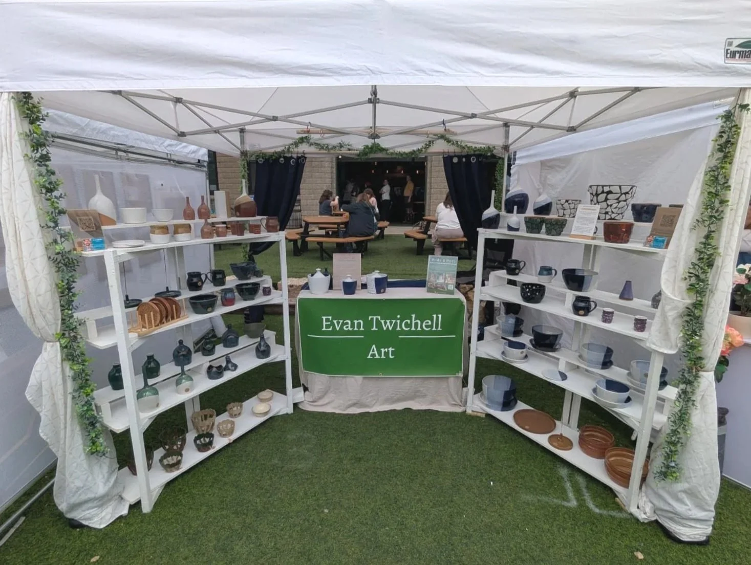 A man in green t-shirt and shorts standing at a vendor booth with ceramic and pottery items and a blackboard sign that reads "Evan Twichell Ceramics" at the Powderhorn Art Fair.