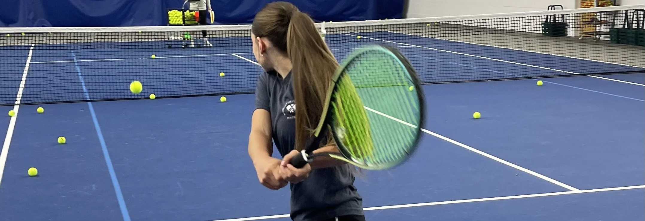 A woman holding a tennis racket on an indoor tennis court, with tennis balls scattered around.