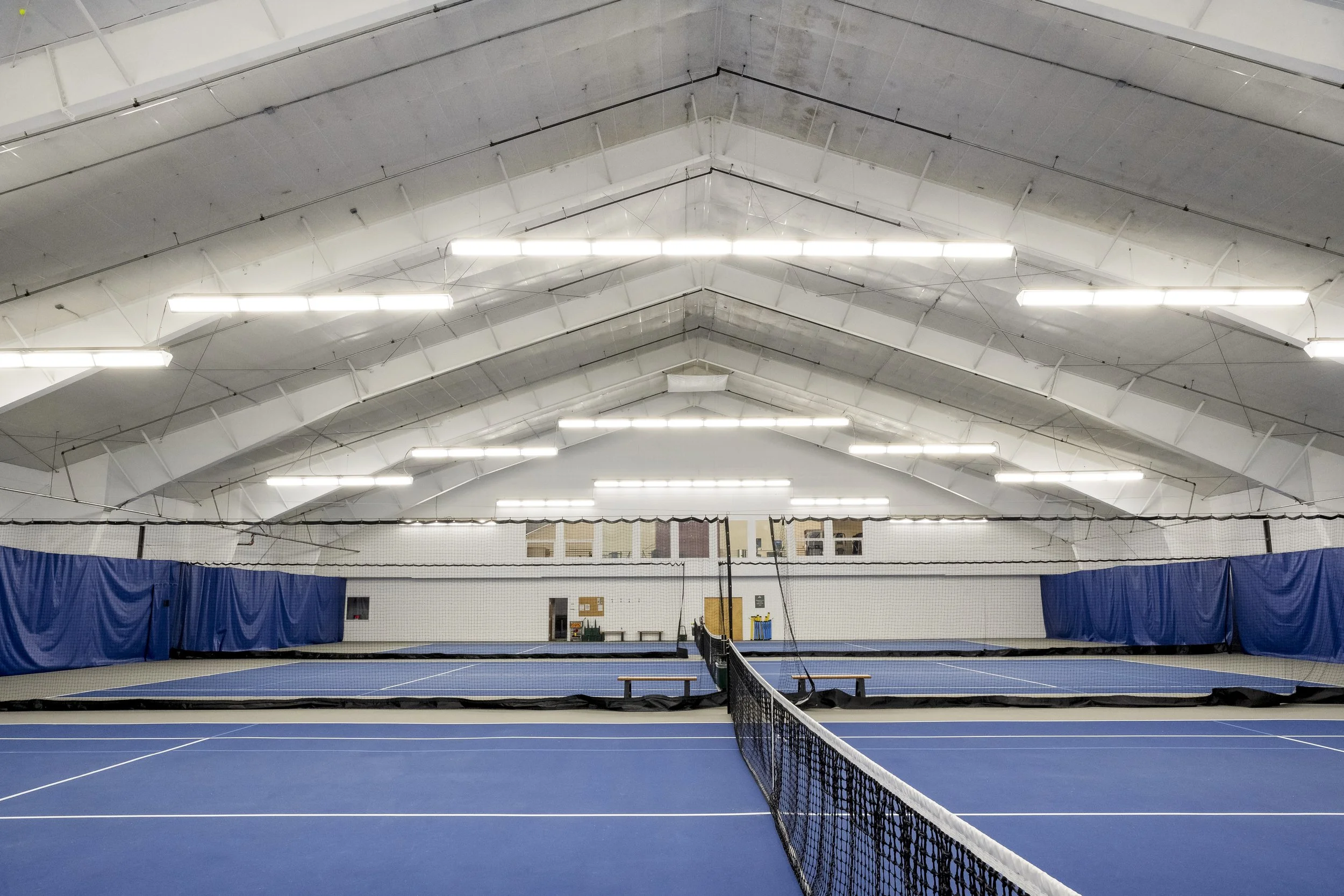Indoor tennis courts with blue courts, net, overhead lighting, and blue curtains along the sides.