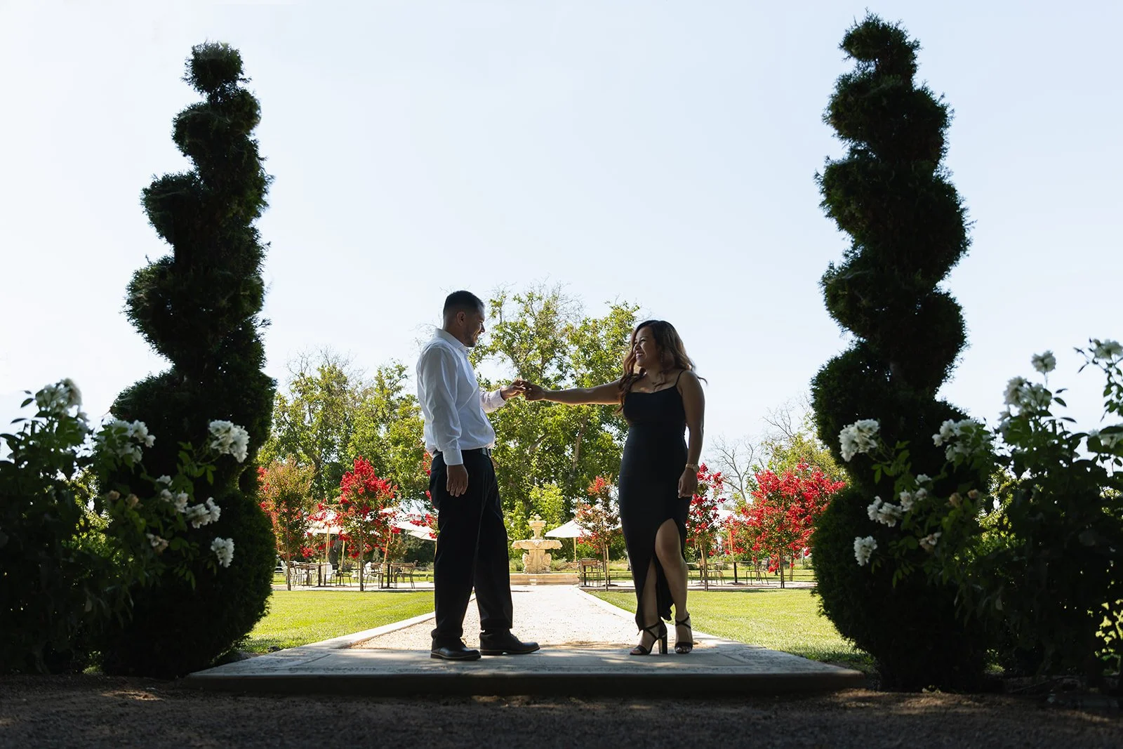 Couple holds hands and dance in front of small water fountain. They are surrounded by tall green bushes and red flower trees in the back at Viaggio Winery for their engagement photo session
