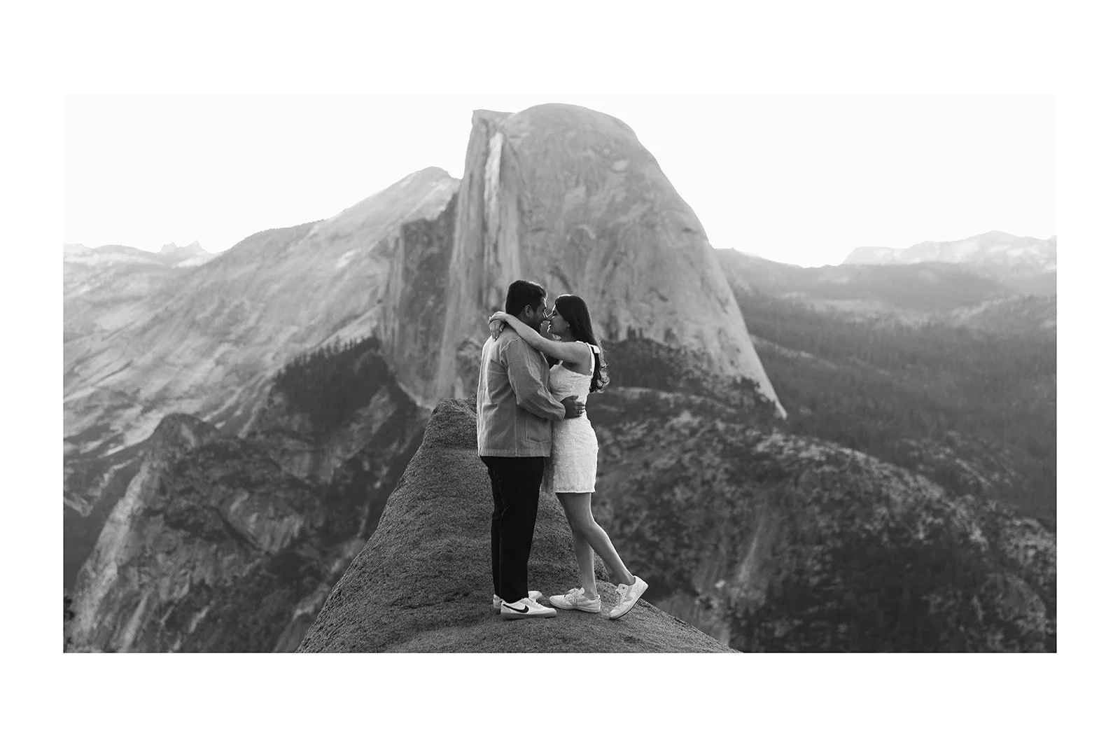 wide landscape engagement photo with couple in small frame standing against backdrop of half dome granite and sky
