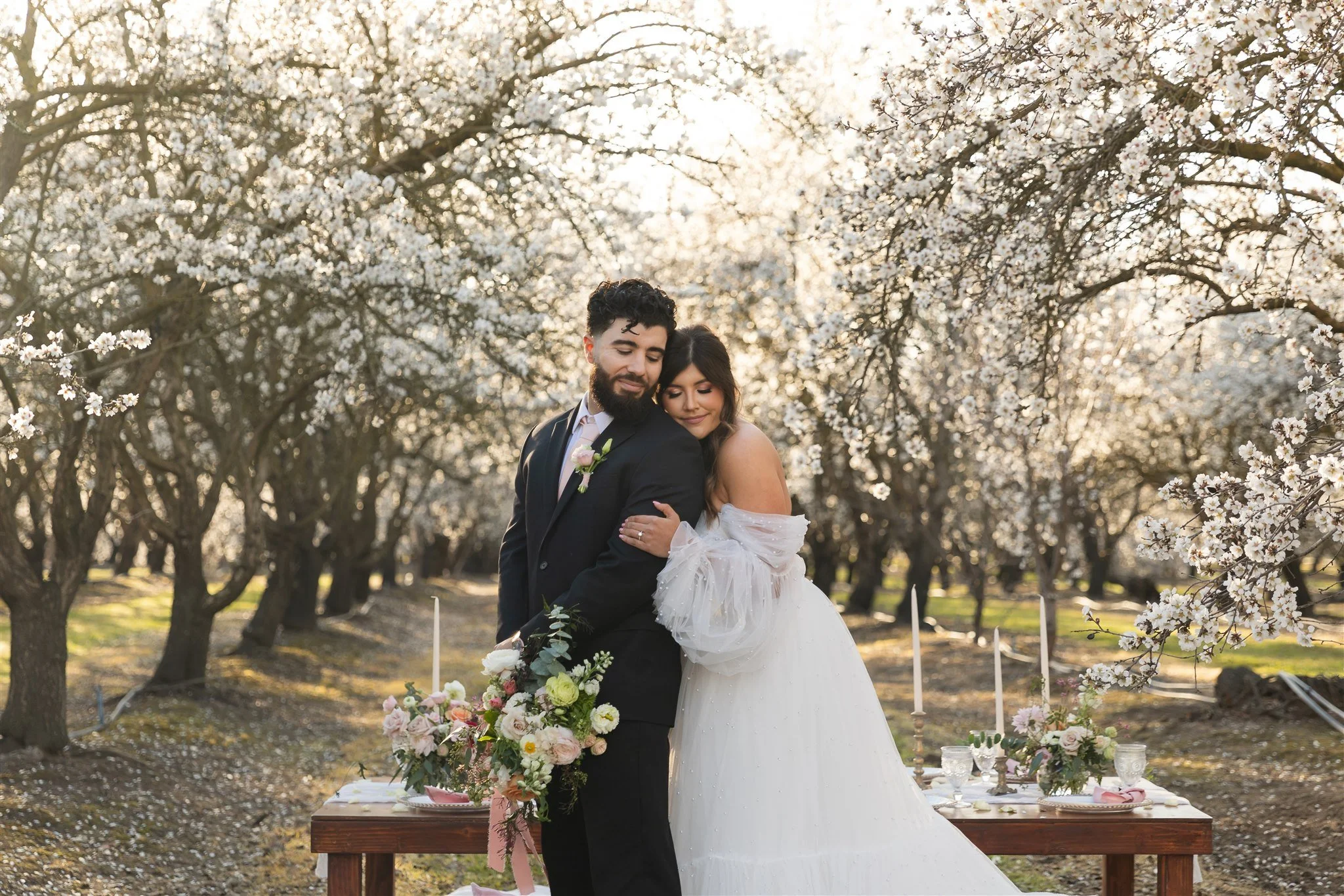 couple embraces in a sweet hug as they are surrounded by warm-light almond blossom trees in Merced California