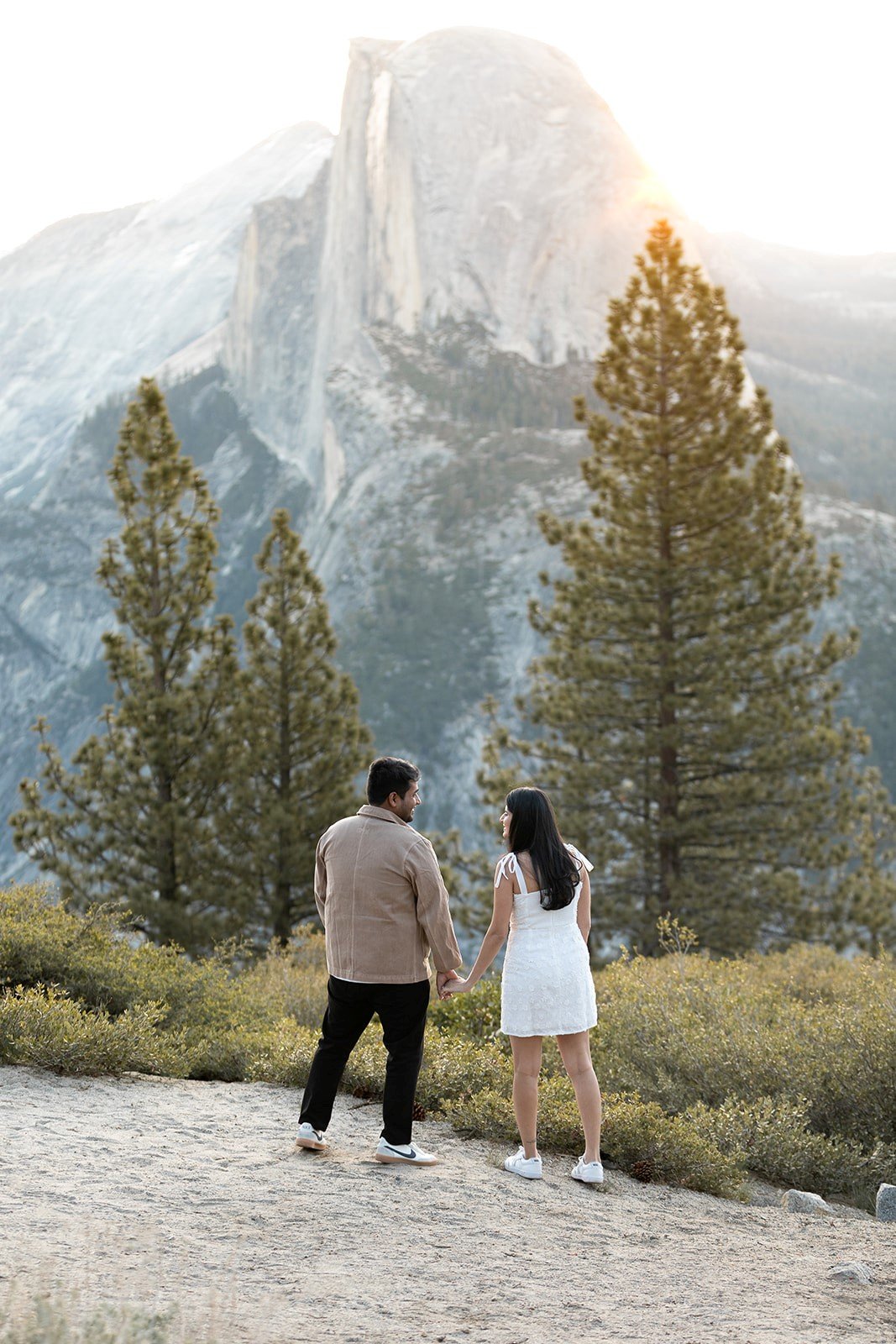 sunrise session of partners holding hands and looking at each other with half dome and 3 large pine trees in slightly blurred background while the sun lights up on the right side of the mountains