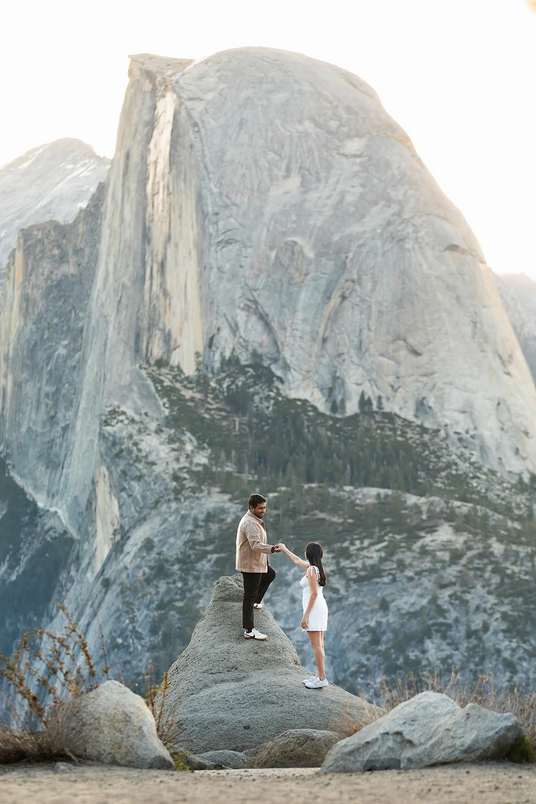 couple standing in rock at glacier point with large backdrop of half dome as the sun peeks from the right side during sunrise