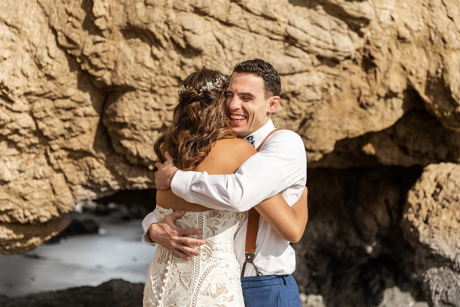 A couple just married and they hug happily after their first kiss. They stand in front of a rock formation on a Malibu beach