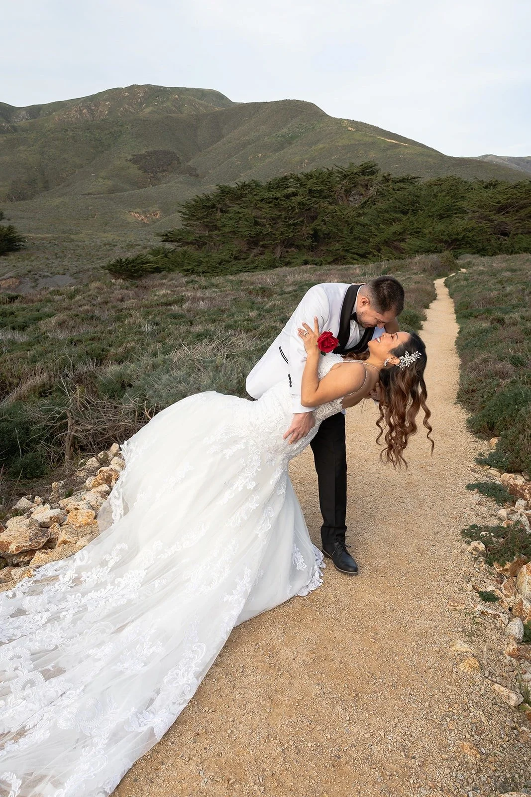 Married couple enjoys a day out in Big Sur weeks after their ceremony, they wear their wedding attire and carry a single red rose